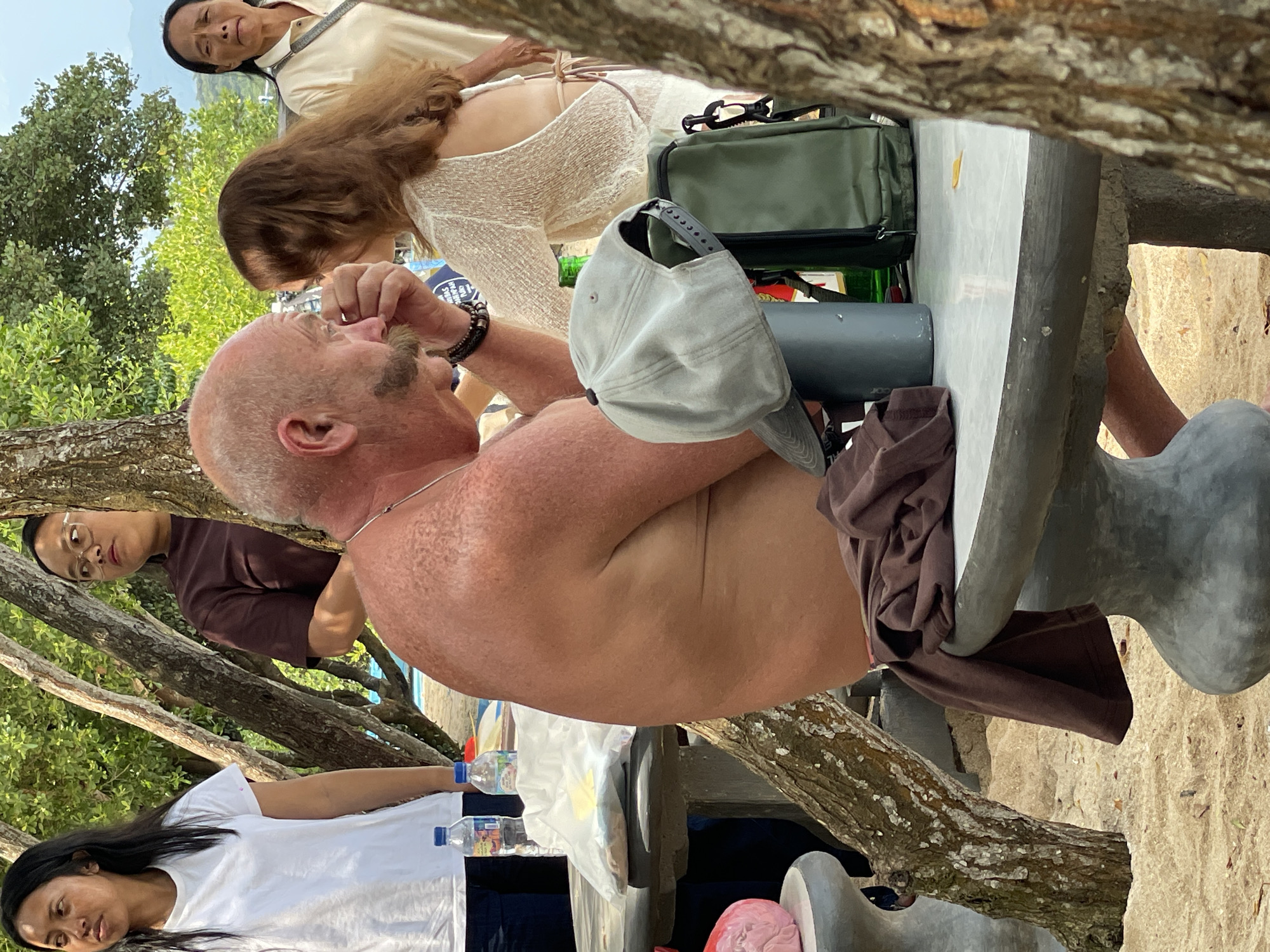 The image depicts an outdoor scene in Gerokgak, Indonesia, featuring five individuals around concrete picnic tables on sandy ground, with prominent tree trunks and lush foliage. In the foreground-center, a bald, middle-aged man with a large handlebar mustache, bare-chested with tanned and freckled skin, is seated at a grey concrete table. He faces a woman with long brown hair, wearing a light-colored, open-back top, whose right hand is positioned near his face. On the table beside them are a grey baseball cap, a dark grey cylindrical container, a green bottle, and a dark green rectangular bag. A brown t-shirt is draped over the concrete stool next to the man. To the left, a young woman with long dark hair, wearing a white t-shirt and dark pants, stands beside another concrete table holding two plastic water bottles and a white plastic bag. Behind her, partially obscured by a tree, another young woman with glasses and a dark brown t-shirt looks towards the right. In the right background, an older woman with dark hair, wearing a light-colored polo shirt, stands observing the scene. The background consists of dense green trees and distant mountains under a bright sky.