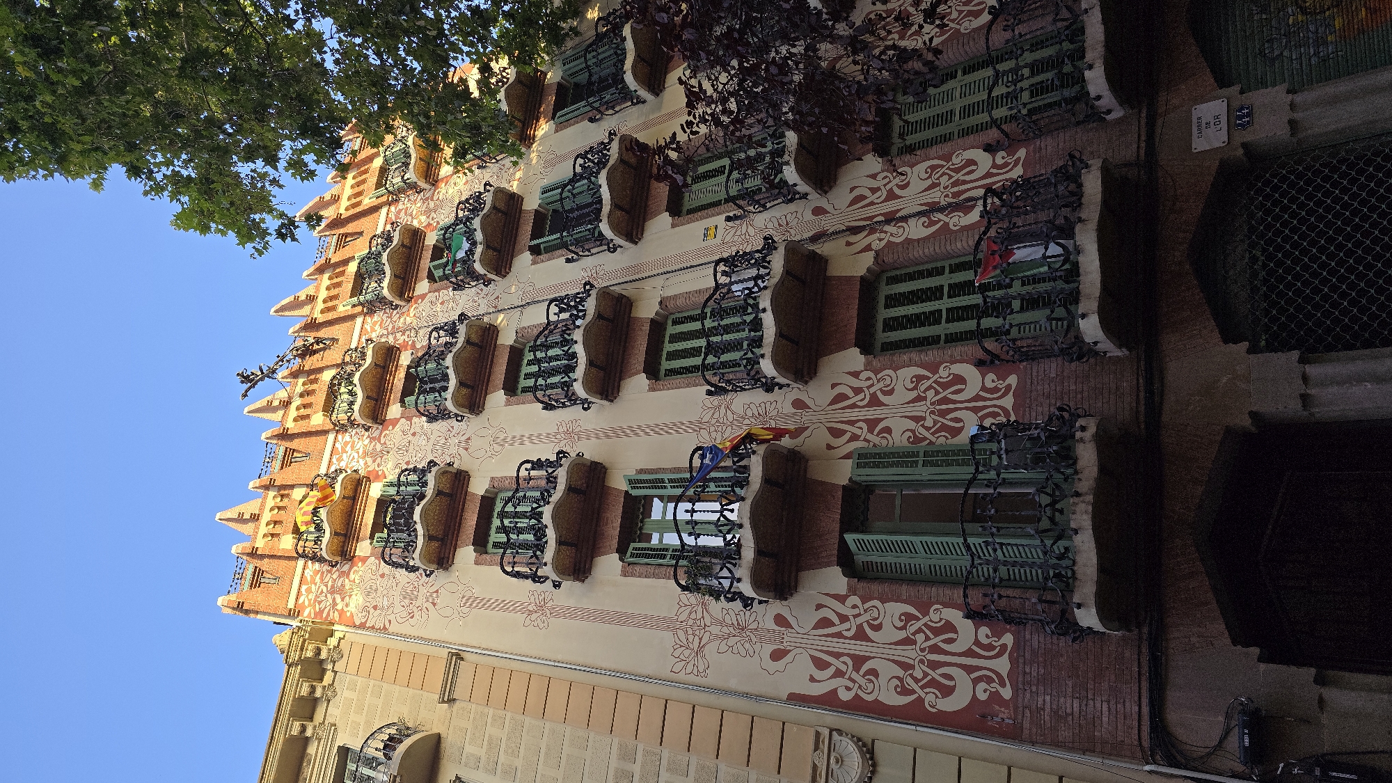 The image depicts the facade of a building in Barcelona, Spain, featuring a distinctive architectural style with ornate balconies and decorative patterns. The building is tall, with multiple floors, and its facade is a mix of light-colored plaster with intricate red and off-white floral or abstract motifs. The balconies are made of wrought iron and have curved, scalloped fronts, with decorative railings. The windows have green shutters, many of which are partially or fully open.
Several balconies display flags. On the upper floors, one balcony has a yellow and red striped flag, likely the flag of Catalonia. Another balcony on a lower floor has a flag that appears to be the flag of Palestine.
The building's roofline is jagged and decorative, made of brick. A large green tree with dense foliage is visible on the right side of the image, partially obscuring the upper right portion of the building. The sky above is a clear, bright blue.
At the base of the building, an arched entrance is visible, with a dark, recessed doorway. To the right of the entrance, a street sign indicates "CARRER DE L'OR" (Street of Gold) with the number "44". Electrical conduits and boxes are attached to the wall near the ground level. The overall impression is one of a well-maintained, architecturally significant building in an urban setting.