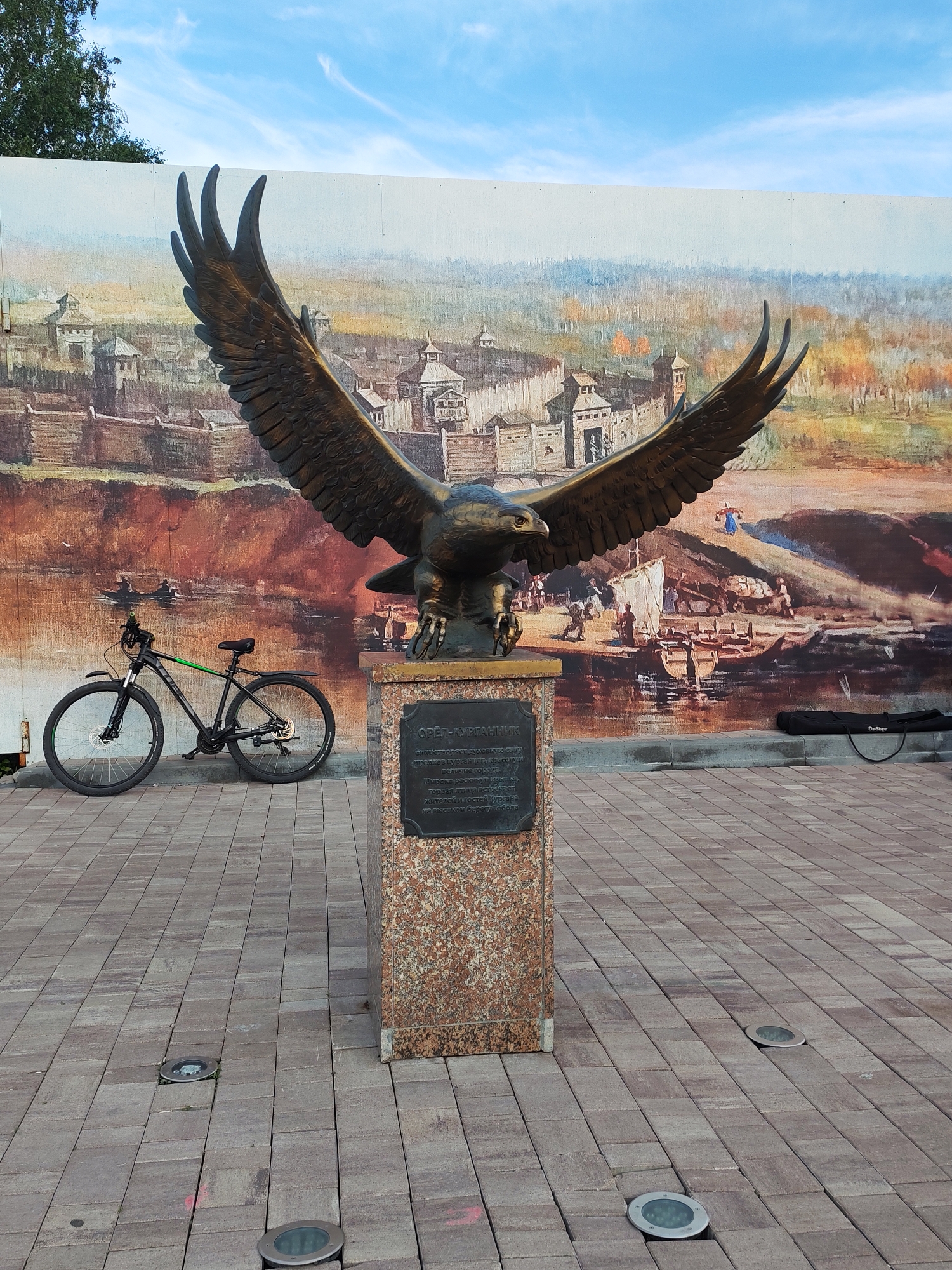 A bronze eagle statue with outstretched wings stands on a granite pedestal in front of a large mural depicting an ancient settlement. To the left of the statue, a black bicycle with a green stripe on its frame is parked on a paved walkway made of interlocking brick-like pavers. The mural shows wooden fortifications, boats on a river, and people engaged in various activities. The sky above is blue with scattered white clouds. The scene appears to be outdoors, possibly in a public square or park. The eagle statue is a prominent landmark in the city of Kurgan, Russia, specifically the "Eagle-Kurganets" monument, commemorating the city's founding.