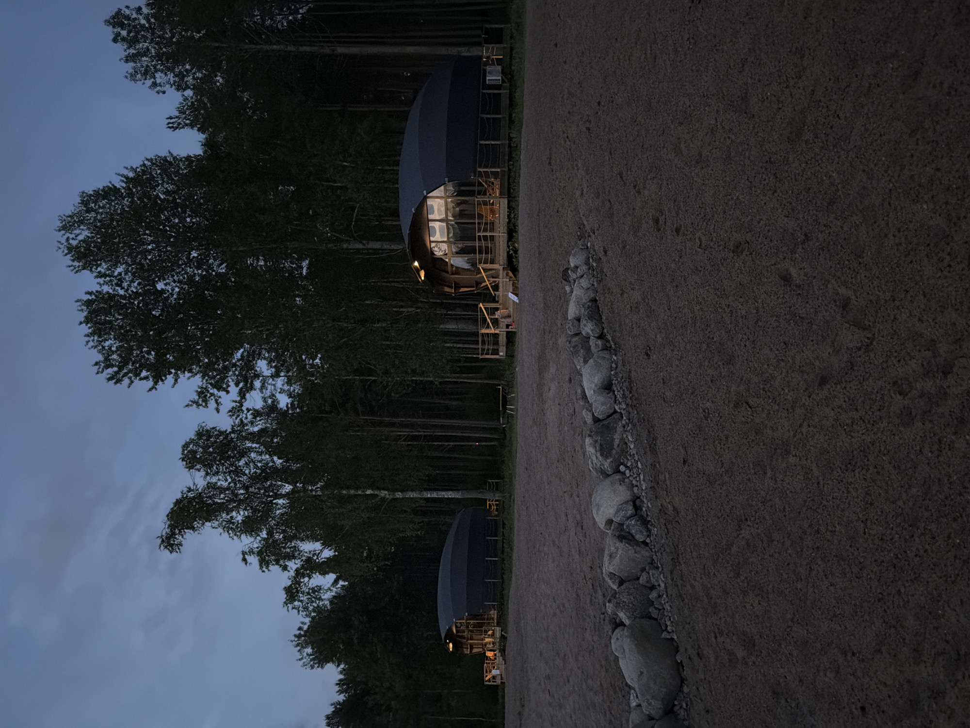 The image depicts a twilight scene in a natural environment, likely a campground or resort, with two dome-shaped structures visible in the distance. The foreground is a sandy area with a row of large stones along its edge. The structures, which appear to be accommodations, are situated on elevated wooden platforms and are surrounded by a dense forest of tall trees. The sky is dark and overcast, indicating either early morning or late evening. Lights are visible inside the structures, suggesting occupancy. No people are directly visible in the image. The location is identified as Solov'evo, Russia.