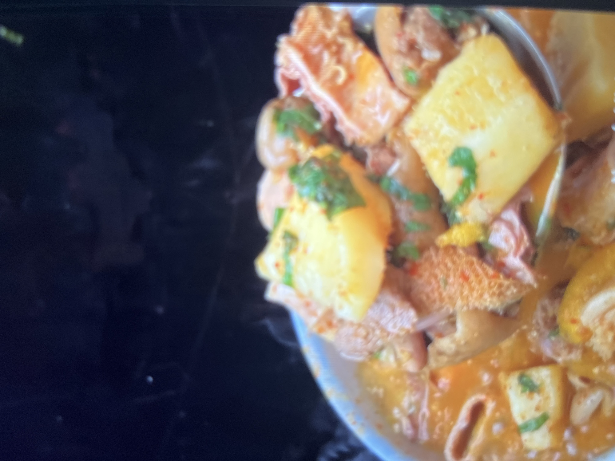 The image shows a close-up of a bowl of food, likely a stew or soup, being served with a spoon. The food appears to contain pieces of meat or tripe, along with large chunks of what seem to be potatoes or yams, and chopped green herbs. The broth is a reddish-orange color, suggesting it may be spiced. Steam is visible rising from the dish, indicating it is hot. The background is dark and out of focus, with some faint lights. There are no people or discernible environmental details that would specifically place this image in Abuja, Nigeria.