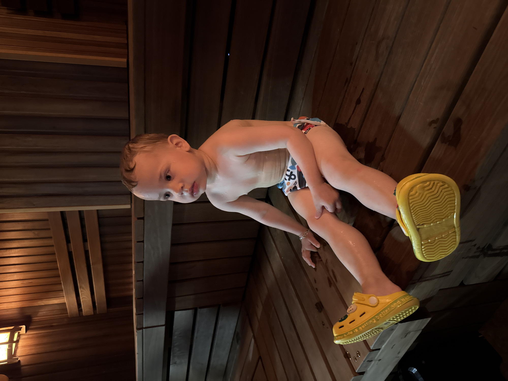 A young, light-skinned child wearing a patterned swim diaper and yellow Croc-style shoes is inside a sauna. The child's skin appears damp. The sauna walls are constructed of wooden planks. A light fixture is visible in the lower left corner. The image does not confirm if the location is in Rusanovo, Russia.