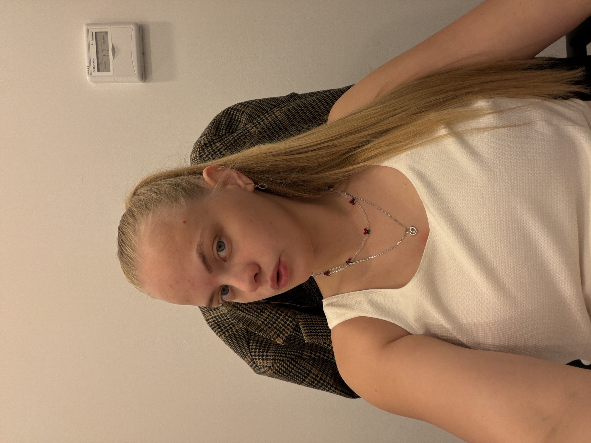 The image shows a woman with long blonde hair and fair skin. She is wearing a white textured tank top and a silver necklace with red beads. She has light blue eyes and is looking towards the camera. She is wearing a brown plaid jacket. The background is a plain white wall. A white digital thermostat is affixed to the wall. It's impossible to determine the specific location in Izhevsk, Russia from this image alone. There are no identifying markers.