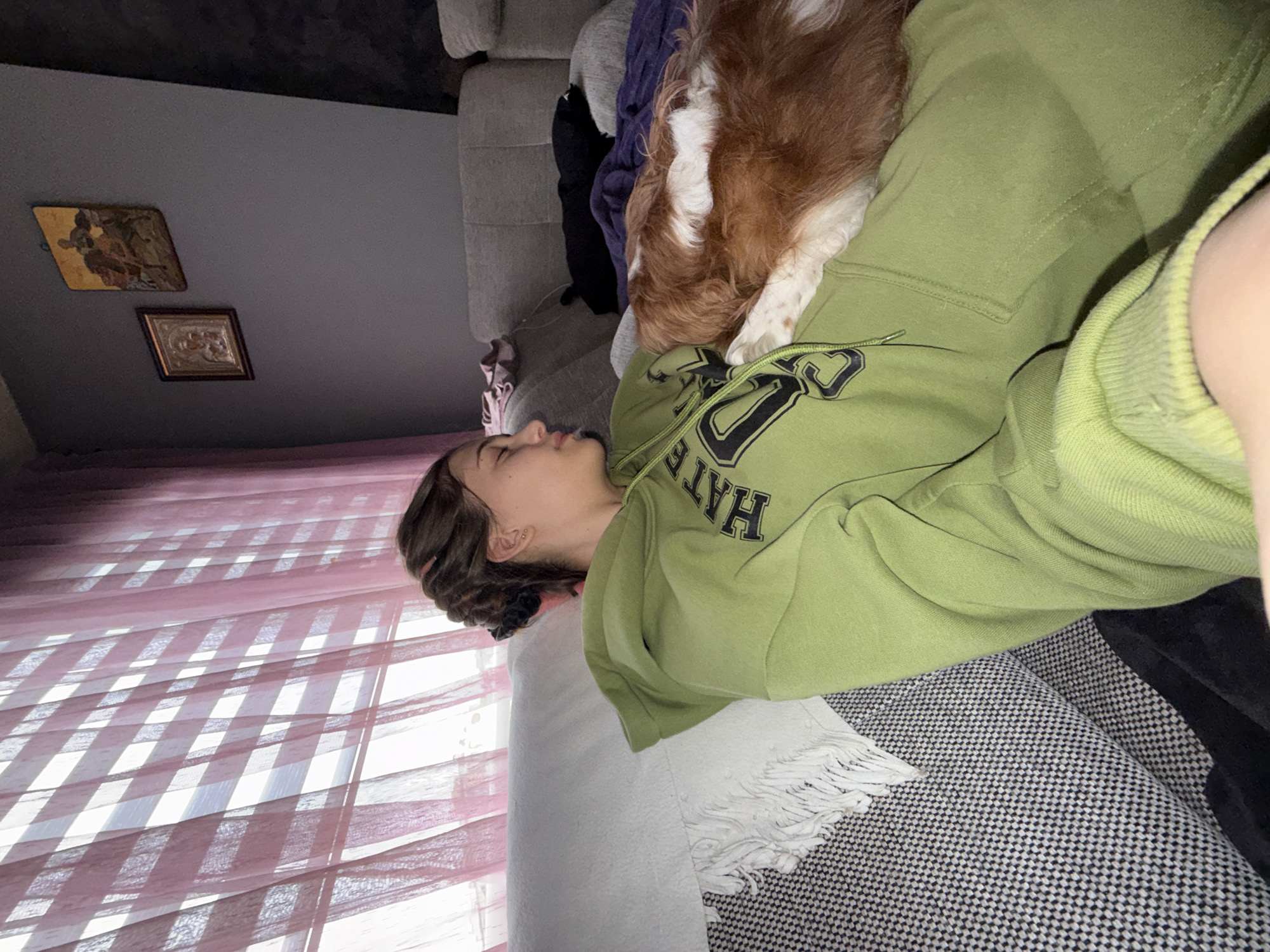 A young woman is lying on a couch in what appears to be a living room. She is wearing a green sweatshirt with black lettering. A brown and white dog is lying on top of her. Behind her is a window with pink curtains. On the wall are two framed pictures. The couch has a gray and white checkered pattern, with a white blanket with fringe. The location is presumed to be in Sofia, Bulgaria, though there is no information in the image to confirm this.