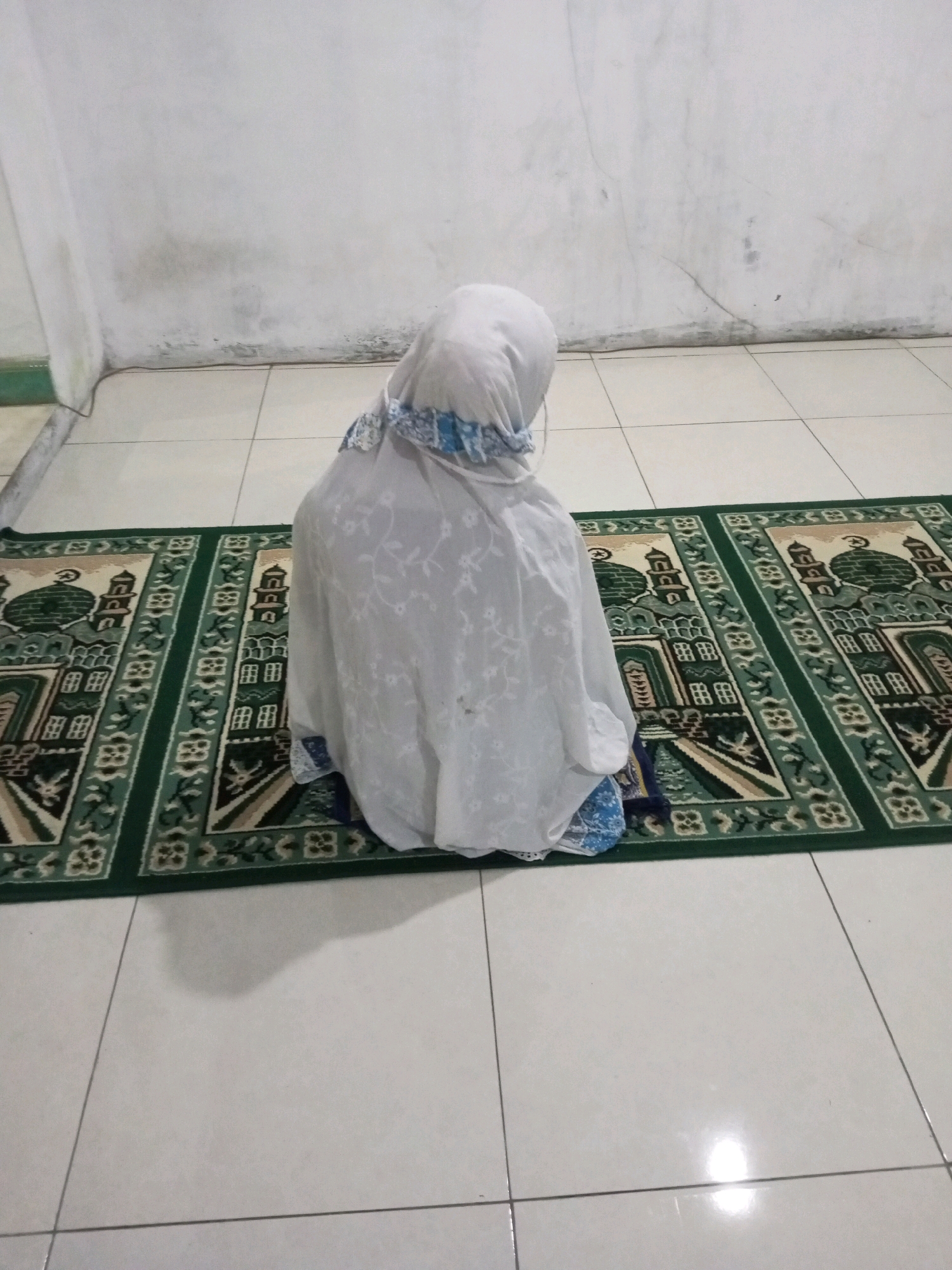 A person wearing a white hijab and a white garment sits on a green prayer rug, facing away from the camera. The rug has a pattern of mosque domes and intricate designs. The person is positioned on a tiled floor in front of a plain, white wall with some water stains near the bottom. The scene appears to be indoors.