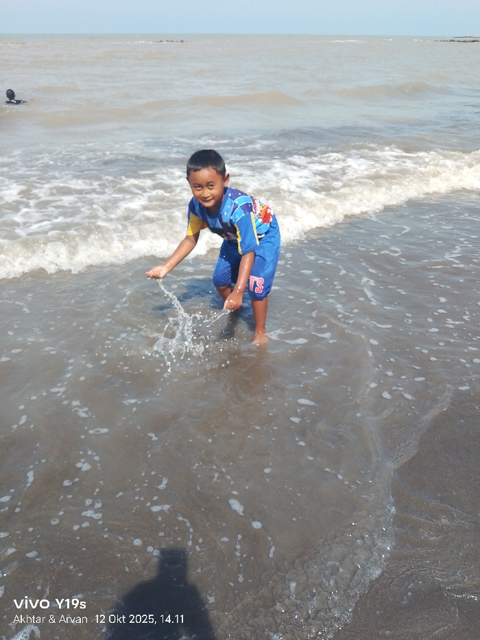 This image captures a beach scene in Limbangan, Indonesia, on October 12, 2025, at 14:11. The primary subject is a young male child, approximately 5-7 years old, standing in shallow, turbid brown ocean water, which reaches just above his ankles. He has short dark hair and is wearing a blue and yellow short-sleeved shirt with various colorful designs, and blue shorts. He is barefoot and actively splashing water upwards with both hands directly in front of him, creating visible water droplets. His face is turned towards the viewer, displaying a smile.
In the far background to the left, the head and shoulders of another person, partially submerged, are visible in the ocean. The ocean water exhibits small, breaking waves with white foam along the shore. The sky above is clear and light blue. A faint, distant landmass or rock formation is discernible on the horizon to the right. The immediate foreground consists of dark, wet sand partially covered by the shallow water. A distinct shadow of a person, likely the photographer holding a device, is cast onto the wet sand and water in the lower-left quadrant of the frame. Metadata "vivo Y19s" and "Akhtar & Arvan" are imprinted on the image.
