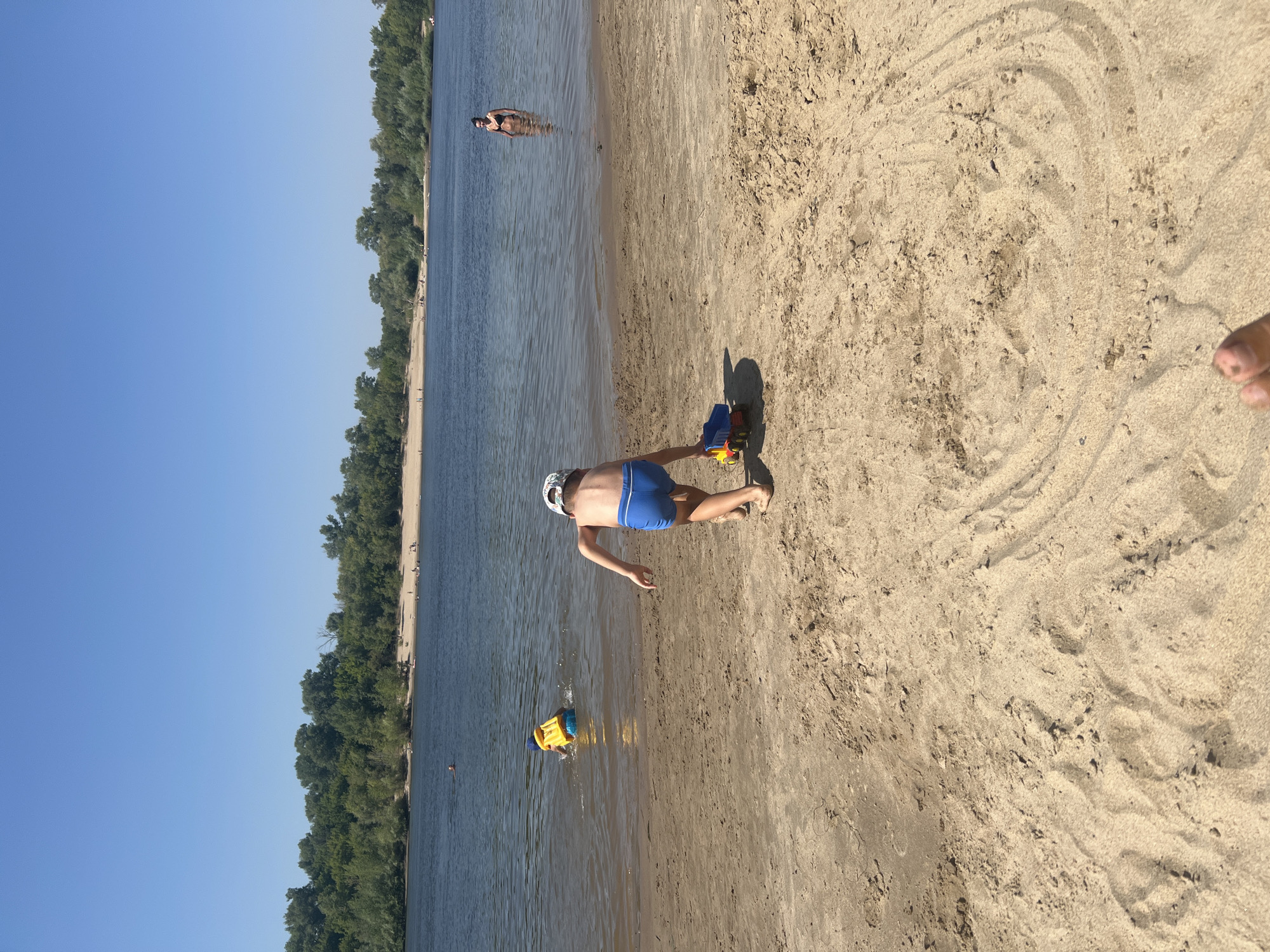 The image shows a sandy beach beside a body of water, likely a river. The sun is bright, indicating it is daytime, and the sky is clear blue. The scene is a recreational area, with several people visible.
In the foreground, a young boy is on the sandy shore, wearing blue swim trunks and a white patterned hat. He is in a push-up position, with a toy truck in front of him. A second child is in the water, playing with a yellow and blue inflatable toy. Further in the water, a woman is swimming or standing. Along the distant shoreline, a line of lush green trees forms a dense treeline. Scattered along the beach and in the water, there appear to be other individuals enjoying the day.
The sandy area shows natural patterns and textures, with some footprints and indentations. The overall mood appears to be relaxed and summery, consistent with a day at the beach. There is no visible text in the image. The location is identified as Kilyakovka, Russia.