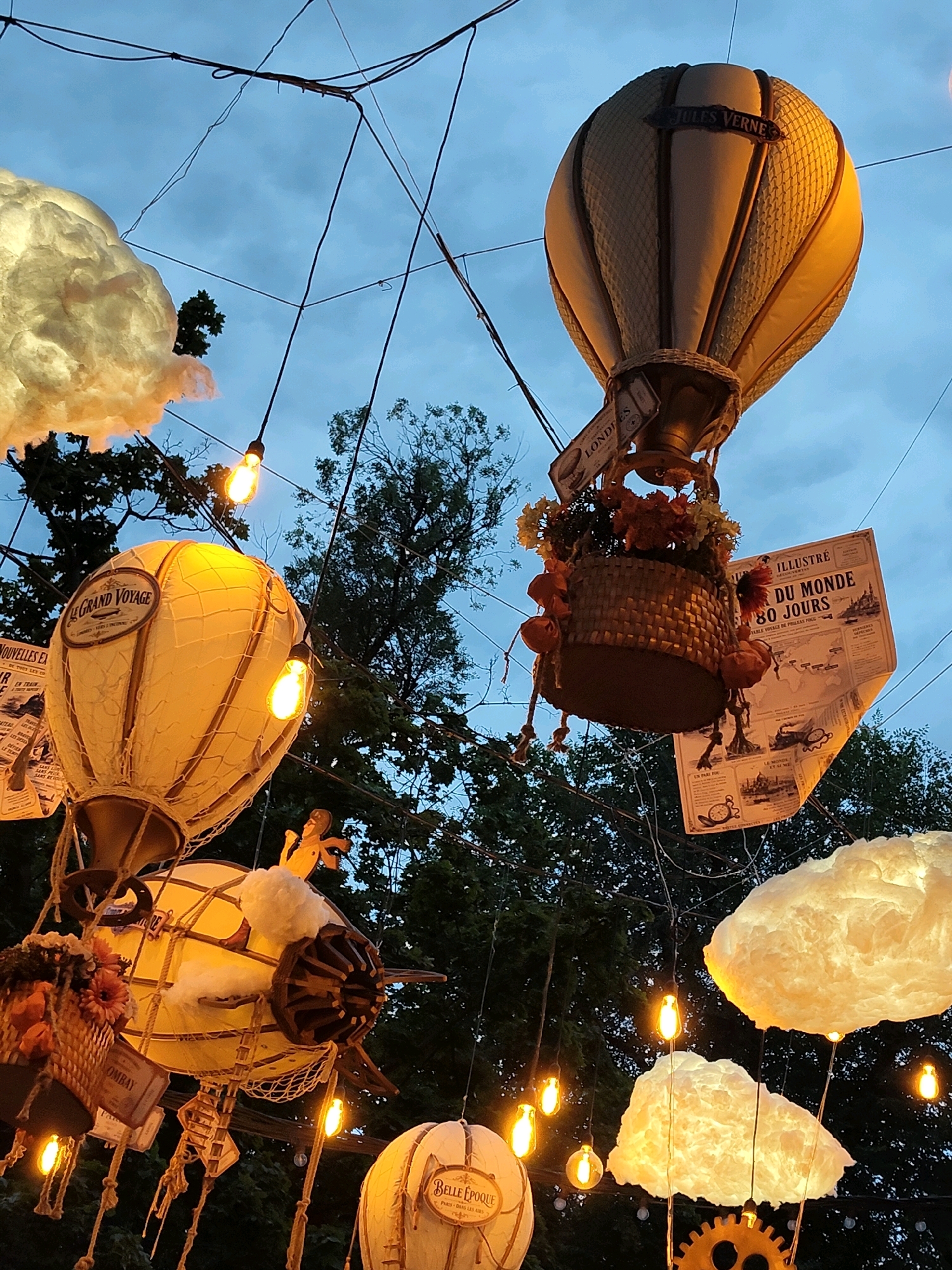 In Chișinău, Moldova, an outdoor display features several decorative hot air balloons suspended by wires against a twilight sky. The balloons are illuminated, creating a warm glow. One prominent balloon is labeled "JULES VERNE" and has a basket filled with artificial flowers. Another balloon, labeled "LE GRAND VOYAGE," is also visible. Scattered among the balloons are illuminated cloud-like decorations and vintage-style light bulbs. A large, illustrated newspaper page titled "ILLUSTRÉ DU MONDE 80 JOURS" is displayed near one of the balloons. The background consists of dark trees and a cloudy sky. The overall scene appears to be a whimsical and festive decoration, possibly for an event or a themed attraction.