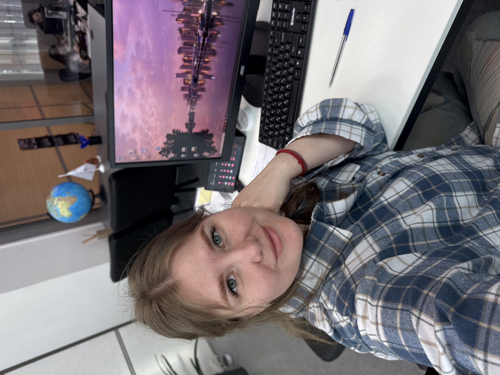 A person is sitting at a desk. A computer monitor displaying an upside-down cityscape with a purple sky is visible. Next to the monitor is a black computer keyboard and a blue pen on a white surface. The person is wearing a blue and white plaid shirt and has a red bracelet on their wrist. A globe is located near the monitor. The location is in Perm, Russia. The presence of a window with blinds indicates the location is indoors, possibly in an office. In the reflection in the window, another person can be seen sitting.