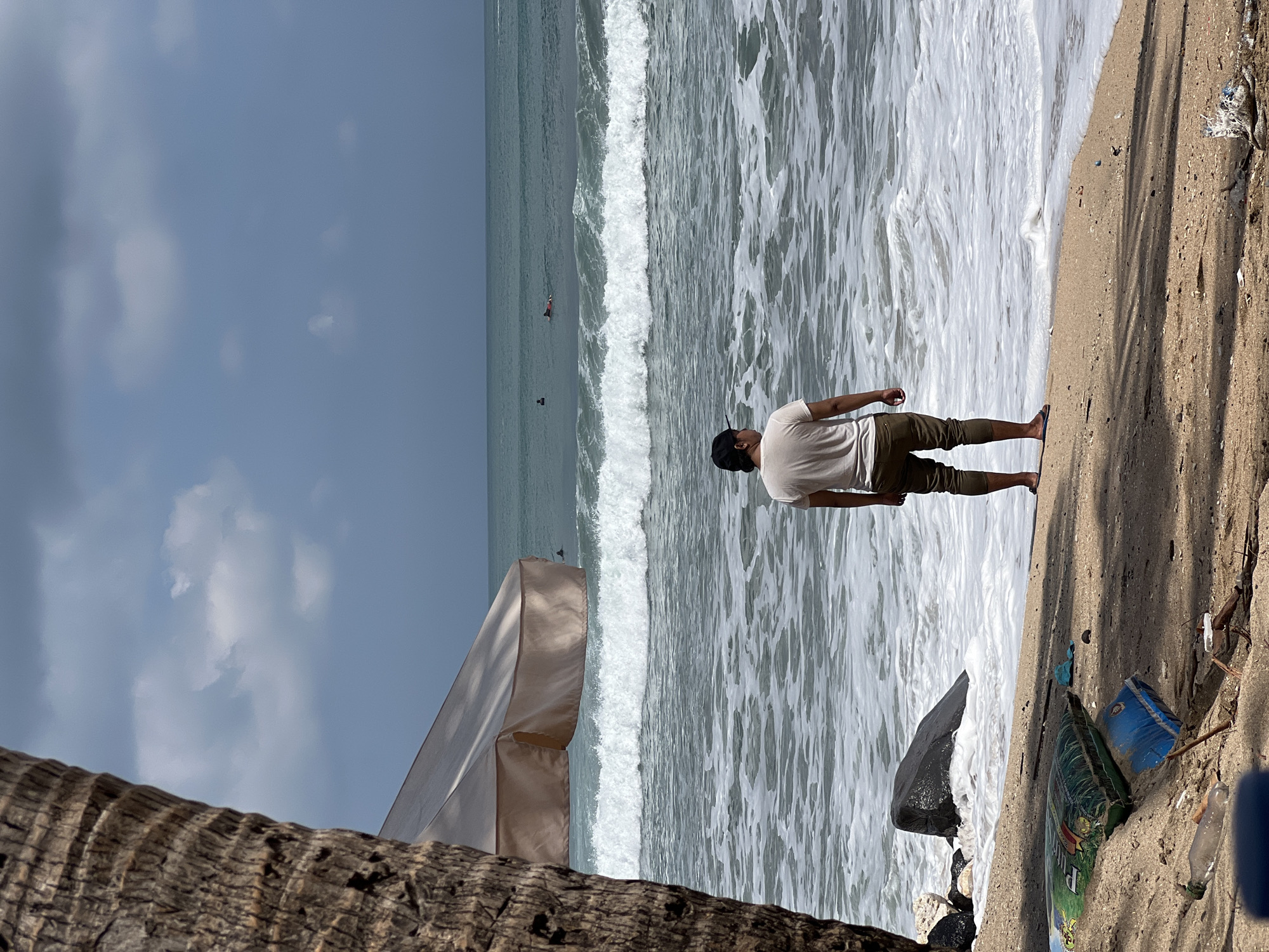 A person stands on the beach in Kuta, Indonesia, facing the ocean. The person is wearing a white t-shirt, green pants, and flip-flops. In the background, waves crash on the shore, and several surfers are visible in the water. A large rock sits near the water's edge. A palm tree trunk is in the foreground on the left, partially obscuring a beach umbrella. Debris, including plastic bags and a bottle, is scattered on the sand. The sky is cloudy.