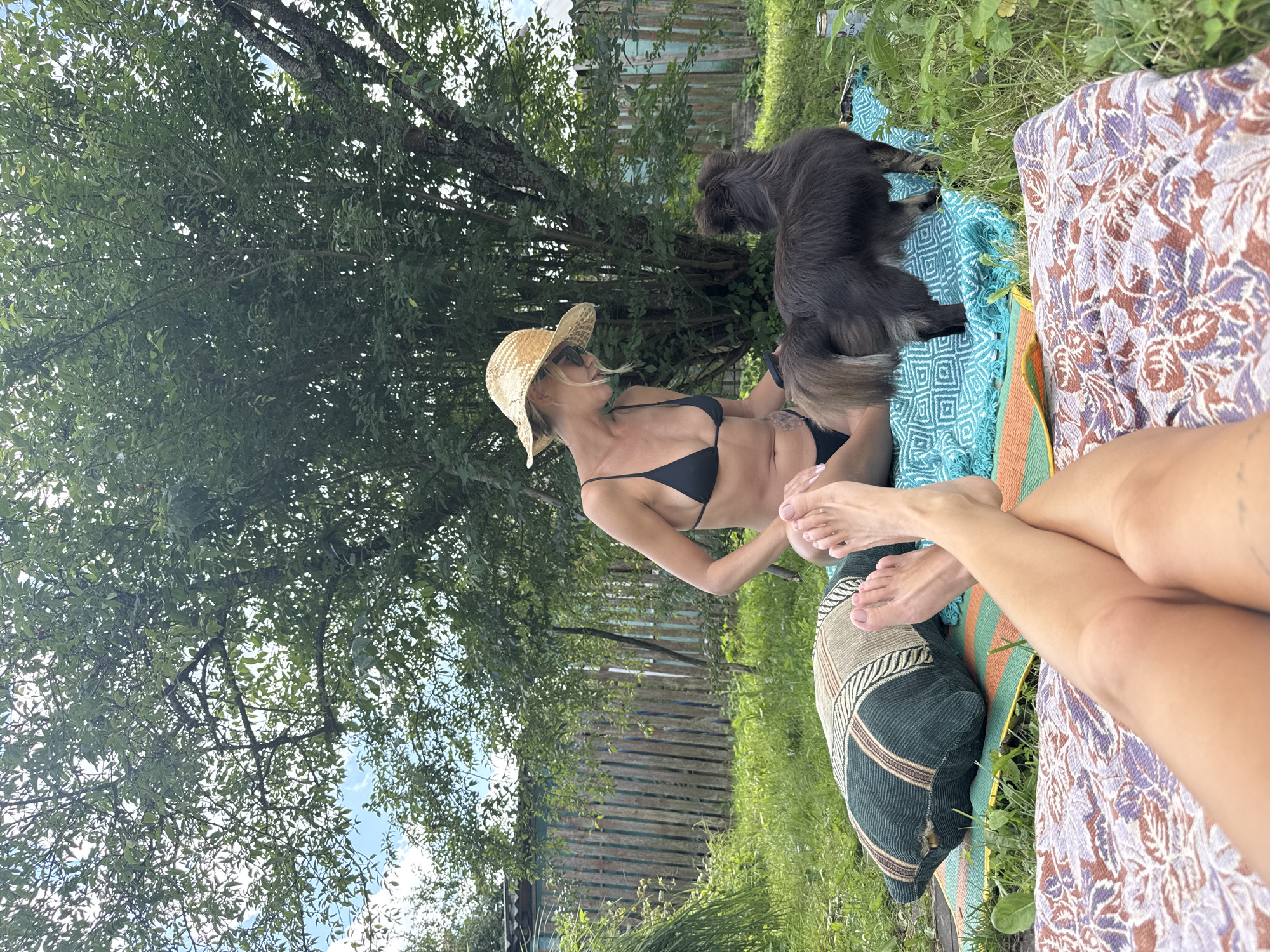 A low-angle outdoor shot captures a woman in a black bikini and a straw hat sitting on a turquoise and orange woven mat in a grassy area. A dark-furred dog stands in front of her, facing away from the camera. The woman is looking down at the dog. Her legs are visible in the foreground, crossed and resting on a patterned blanket. Behind her, a weathered wooden fence and lush green trees fill the background. The scene appears to be a casual outdoor gathering, possibly in a dacha or garden setting in Красная Пресня Снт, Russia.