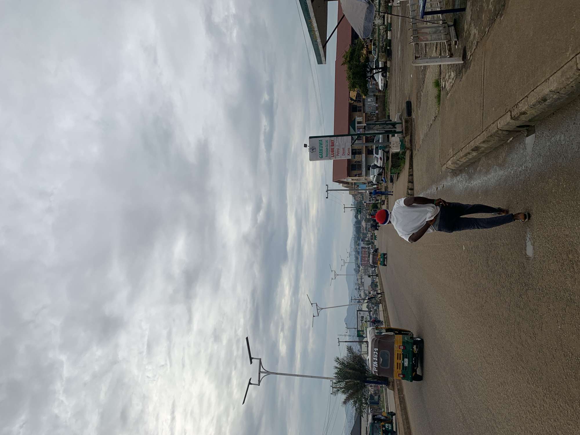 The image captures a street scene in Rafin Pa, Nigeria, under a cloudy sky. The road is paved with asphalt and shows signs of wear. A person wearing a white t-shirt and dark pants, with a red cap, walks away from the camera on the right side of the road. To the left of the road, a yellow and green auto-rickshaw with the license plate "PTA-1634" is moving forward. Several other auto-rickshaws and cars are visible on the road, indicating a moderately busy traffic flow. Along the right side of the road, a sidewalk is present, with a sign for "GARKUWA PETROLEUM Co. Ltd. LUBE BAY" and listings for "Petrol," "Diesel," and "Kero." Further down the road, buildings with red roofs line the street, and distant hills are visible. Streetlights are spaced along the left side of the road. The overall environment suggests an urban or semi-urban setting.