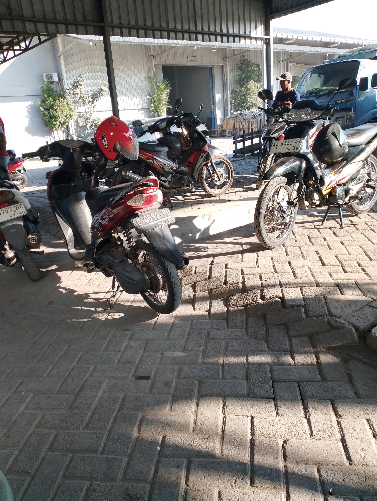 The image depicts an outdoor scene, likely a parking area under a covered structure. Several motorcycles are parked on a paver stone surface. In the foreground, a red helmet rests on the handlebars of a dark-colored scooter. To the right, a black motorcycle with yellow and red accents is parked, its license plate reading "AG 6553 HI 04.29". Behind it, a blue truck is visible. A person wearing a cap and a blue shirt stands near the truck, holding a phone. In the background, a building with corrugated metal walls and a white roof is present, with some vegetation and an air conditioning unit visible on the exterior. The scene appears to be in an industrial or commercial area, possibly in Jambean, Indonesia, given the context provided. The lighting suggests it is daytime, with shadows cast by the structure and the vehicles.