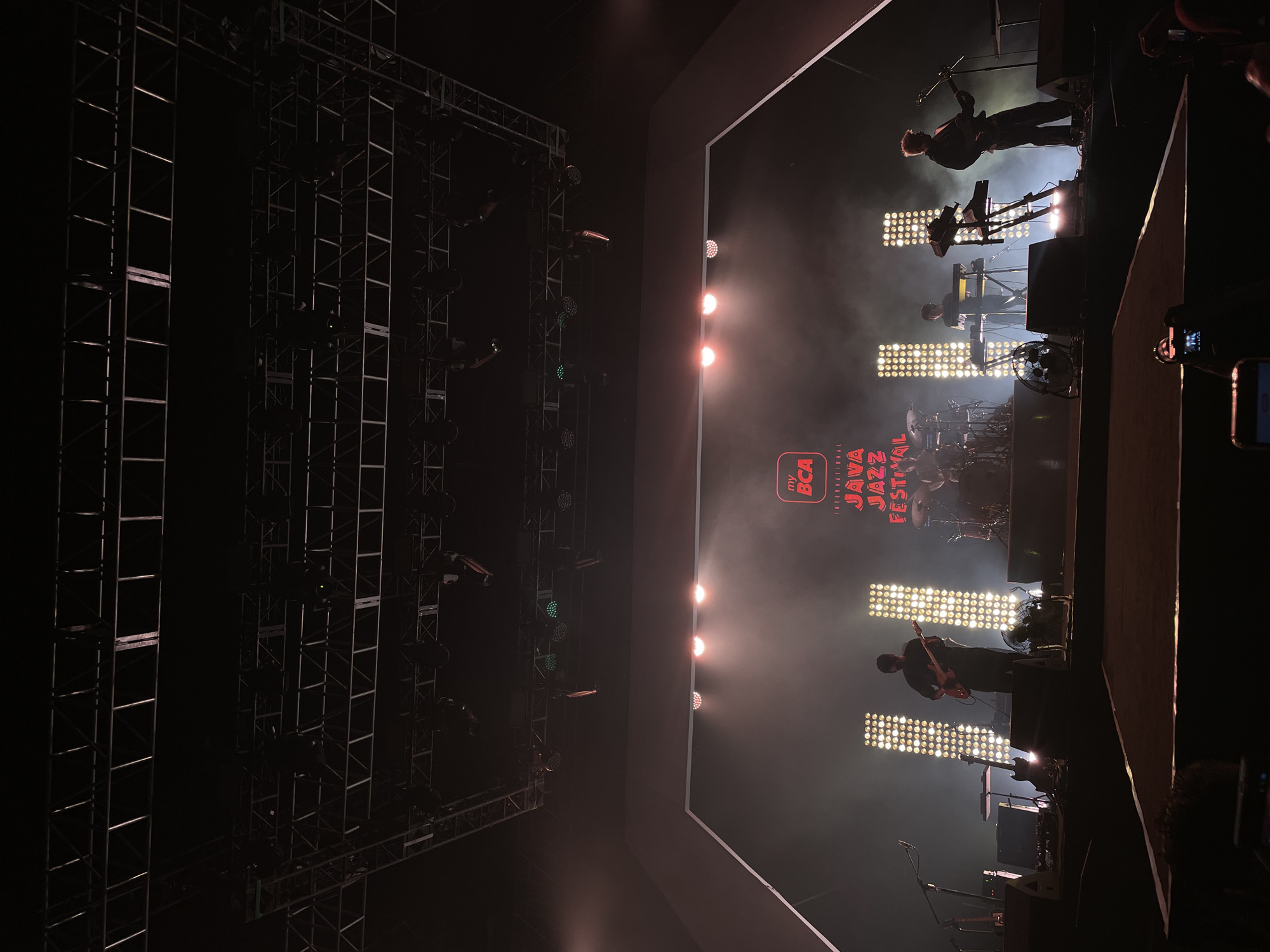 The image shows a band performing on stage at the Java Jazz Festival, likely in Salembaran, Indonesia. The stage is well lit with multiple spotlights. The band members visible include a guitarist, keyboardists, and a drummer. A large metal structure with lighting rigs is visible on the left side of the frame. A logo for the Java Jazz Festival is prominently displayed in the center of the stage. The overall environment is dark, with stage lighting providing the main illumination. Attendees' hands holding phones recording are barely visible in the bottom right.