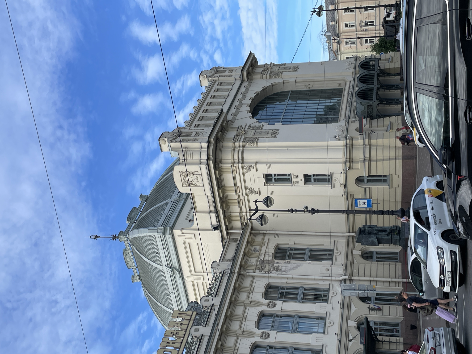 The image shows the exterior of a grand, cream-colored building with a large domed roof, characteristic of Saint Petersburg, Russia. The architecture features ornate detailing, multiple windows, and a prominent arched window on the right side. Several cars, including a white taxi with Russian markings, are visible in the foreground, suggesting an urban street scene. People are seen walking and standing near the building and the vehicles. The sky is blue with scattered white clouds. The sign on the building appears to read "ОКТЯБРЬСКИЙ" which translates to "Oktyabrsky," possibly indicating a station or a public building in Saint Petersburg.
