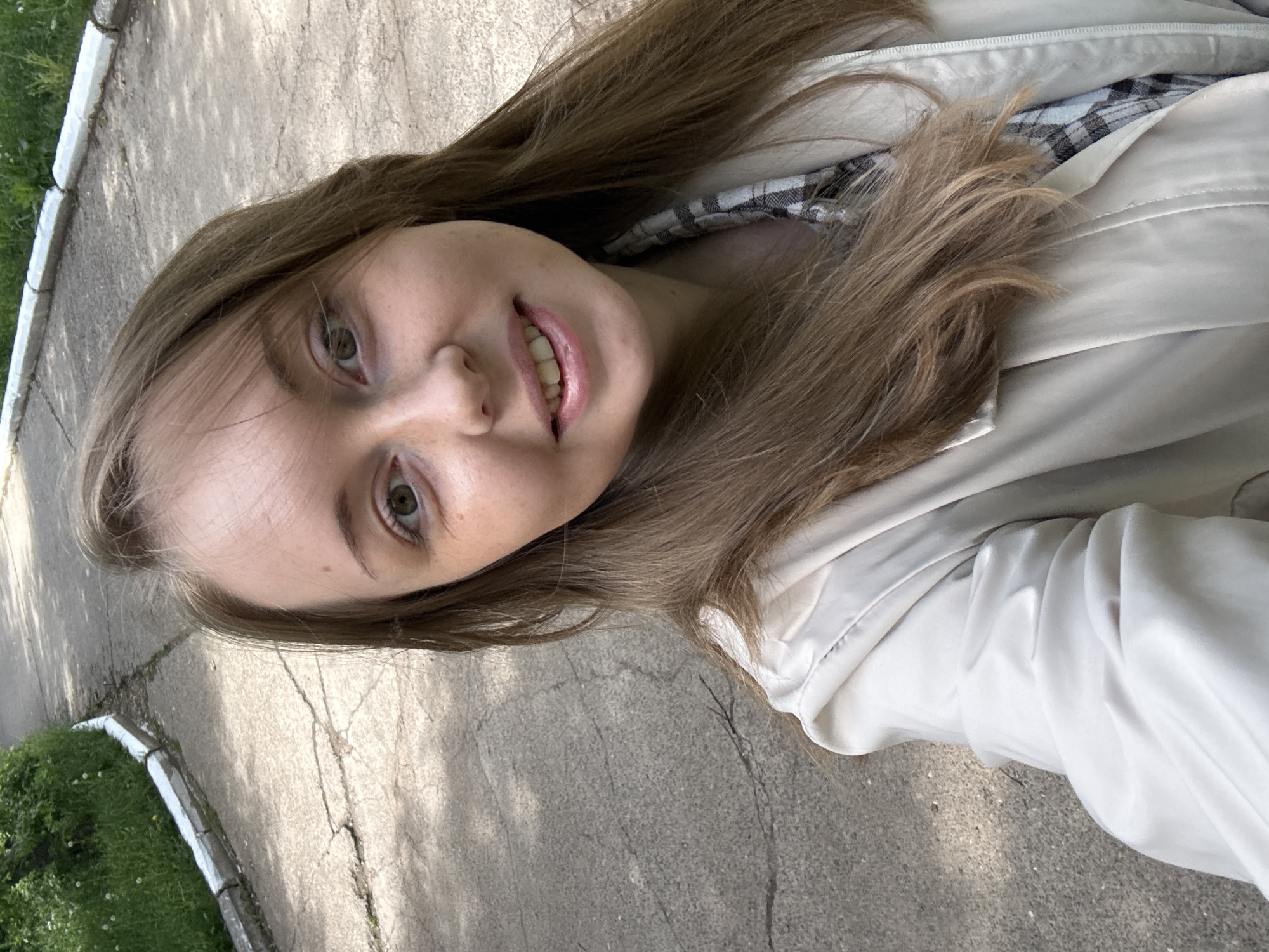 The image shows a young woman with long brown hair and light skin, wearing a white jacket over a plaid shirt. She is smiling slightly and looking directly at the camera. The background appears to be an outdoor setting, possibly a sidewalk with grass and greenery visible in the lower left corner. The location seems to be a city, but the specific details are not clear enough to determine the exact location.