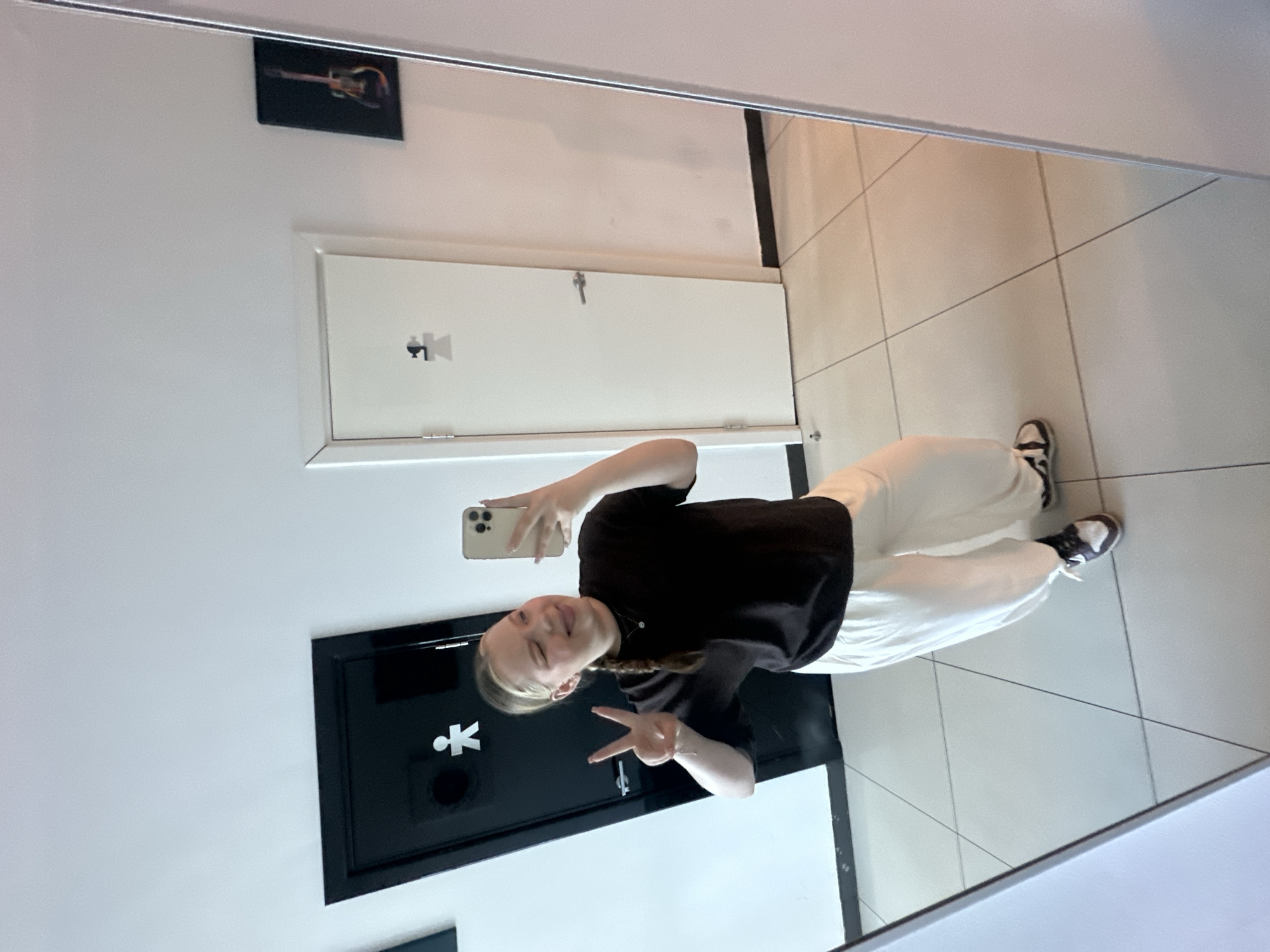 A young woman takes a selfie in a mirror in what appears to be a bathroom. She is wearing a black t-shirt and white pants, with black and white sneakers. She has blonde hair braided down her back and is making a peace sign with her fingers. She is holding a smartphone to take the photo. In the background, the bathroom features white walls with a guitar picture, a white cabinet, and a black door with a stylized "male" restroom symbol. The floor is tiled in a light color. There are no signs to verify the specific location of Chaikovsky, Russia.
