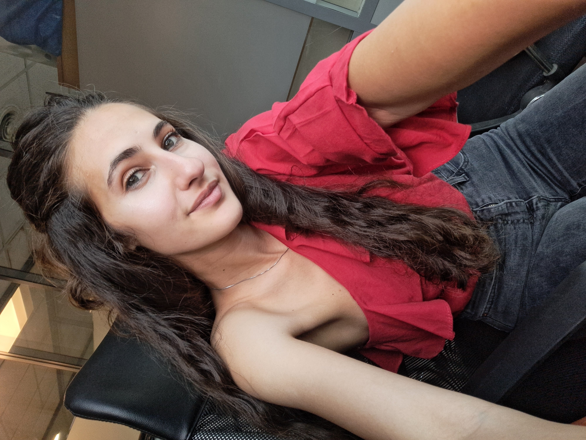 A young woman is lying down on a black office chair. She has long, dark, wavy hair and light skin. She is wearing a red top and grey jeans. She is looking directly at the camera with a slight smile. A thin silver necklace is visible around her neck. The location cannot be determined from the image.