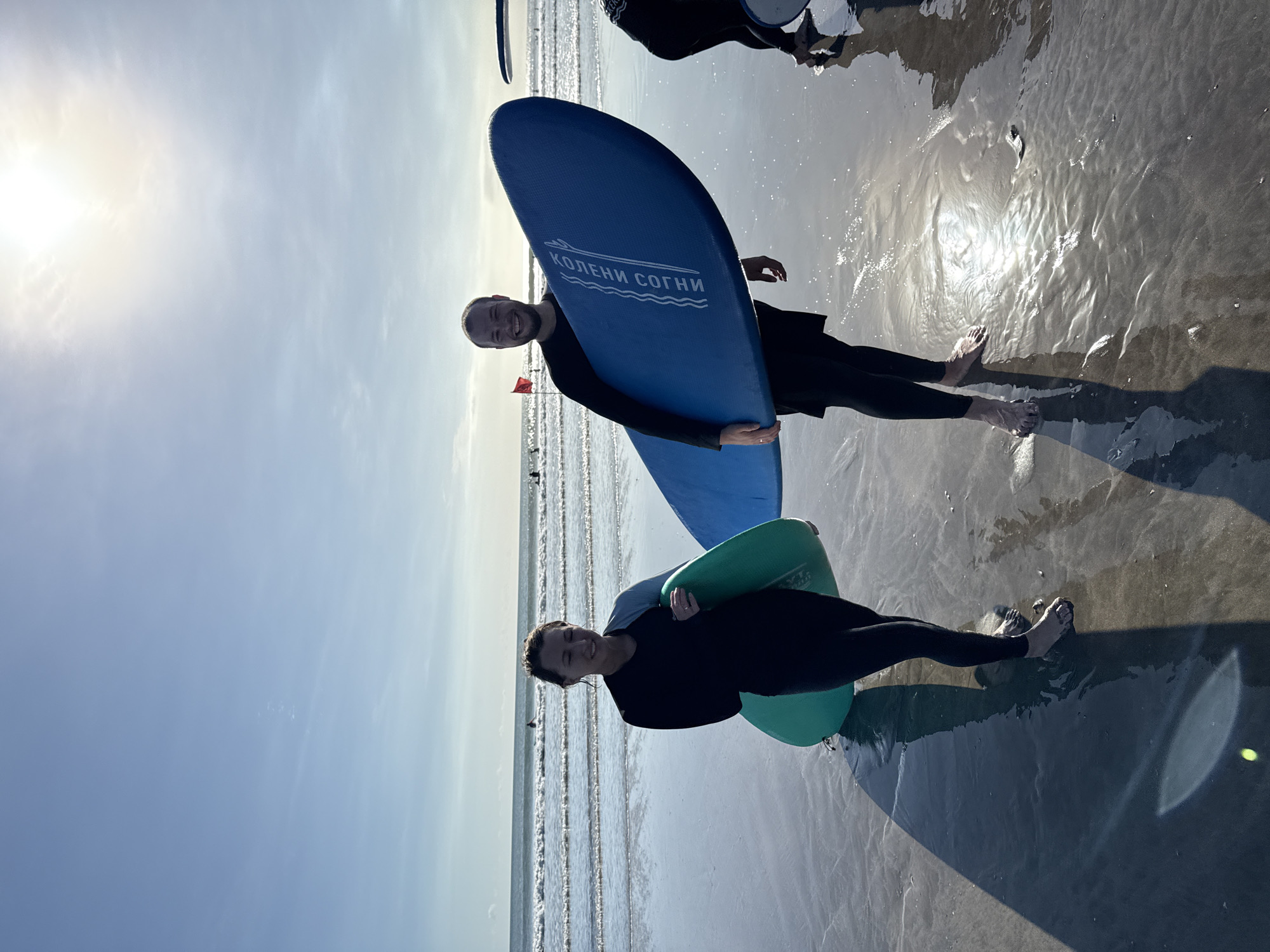Two individuals are standing on a wet, sandy beach, each holding a surfboard. The person on the left has a green surfboard, while the person on the right has a blue one with the inscription "Колени Согни". Both individuals are wearing black wetsuits. The scene is set at Kuta Beach in Indonesia, as suggested by the beach environment and the inscription on the blue surfboard, which is the name of a surfing school operating in Kuta. The sky is bright with a visible sun. The ocean is visible in the background with gentle waves. The scene appears to be captured at a slight angle. Other people are visible in the distance.