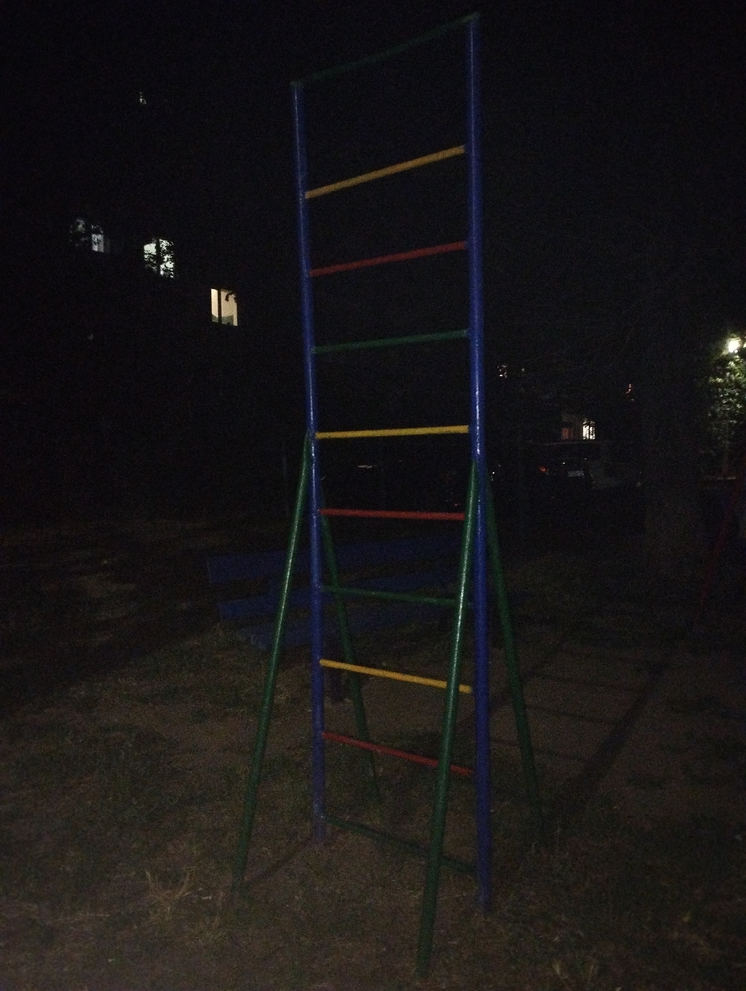 The image shows a nighttime scene in Belaya Kalitva, Russia, featuring a colorful jungle gym with blue, green, red, and yellow bars, set against a dark background. In the distance, several illuminated windows of a building are visible, suggesting a residential area. The ground is covered with dry grass and dirt. The overall atmosphere is dim and quiet, characteristic of a city at night.