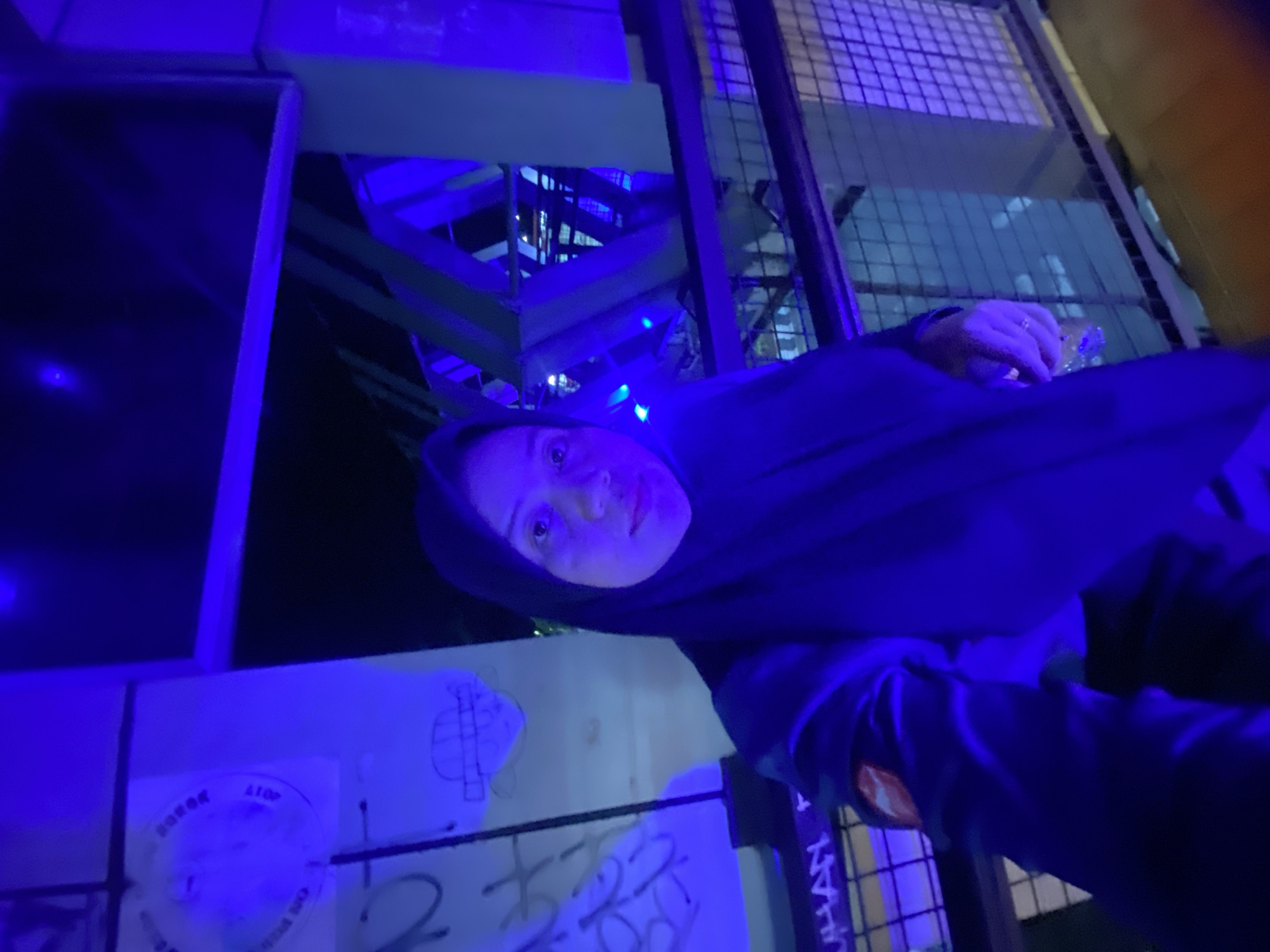 A woman wearing a black hijab and dark clothing takes a selfie in an outdoor setting illuminated by blue lights. She is standing on a walkway with a metal railing in the foreground. In the background, there is a structure with green and blue supports, and graffiti on a white wall. The scene appears to be at the Taman Idaman City Park in Banjarbaru, Indonesia, known for its vibrant blue lighting at night.