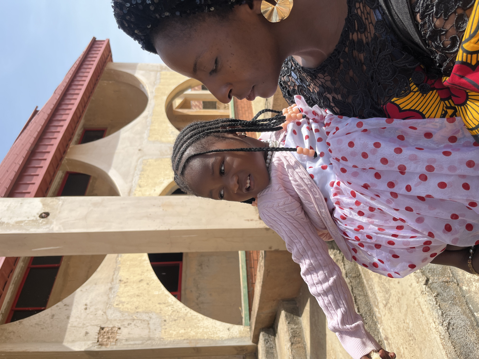 In Bukuru, Nigeria, under a clear sky, a Black woman holds a young Black girl. The woman is on the right, facing left, her profile visible. She has dark skin, dark eyes, and dark hair styled in braids, partially covered by a black sequined headwrap. She wears a black lace top over a brightly patterned garment with yellow, red, and black floral motifs. A large, gold, fan-shaped earring adorns her right ear.
The girl is on the left, facing left and looking upwards. She has dark skin, dark eyes, and her hair is styled in multiple braids adorned with pink beads. She wears a light pink cardigan over a white dress with red polka dots. Her right arm is extended slightly outwards.
They are positioned in front of a building with a distinctive architectural style. The building has a light yellow, textured facade with large, arched openings. The arches are a pale grey concrete, and within them, dark windows are visible. A red, tiled roof with visible rafters caps the structure. To the left of the woman and child, rough, concrete steps lead upwards. The overall setting appears to be outdoors, possibly in a courtyard or public space.