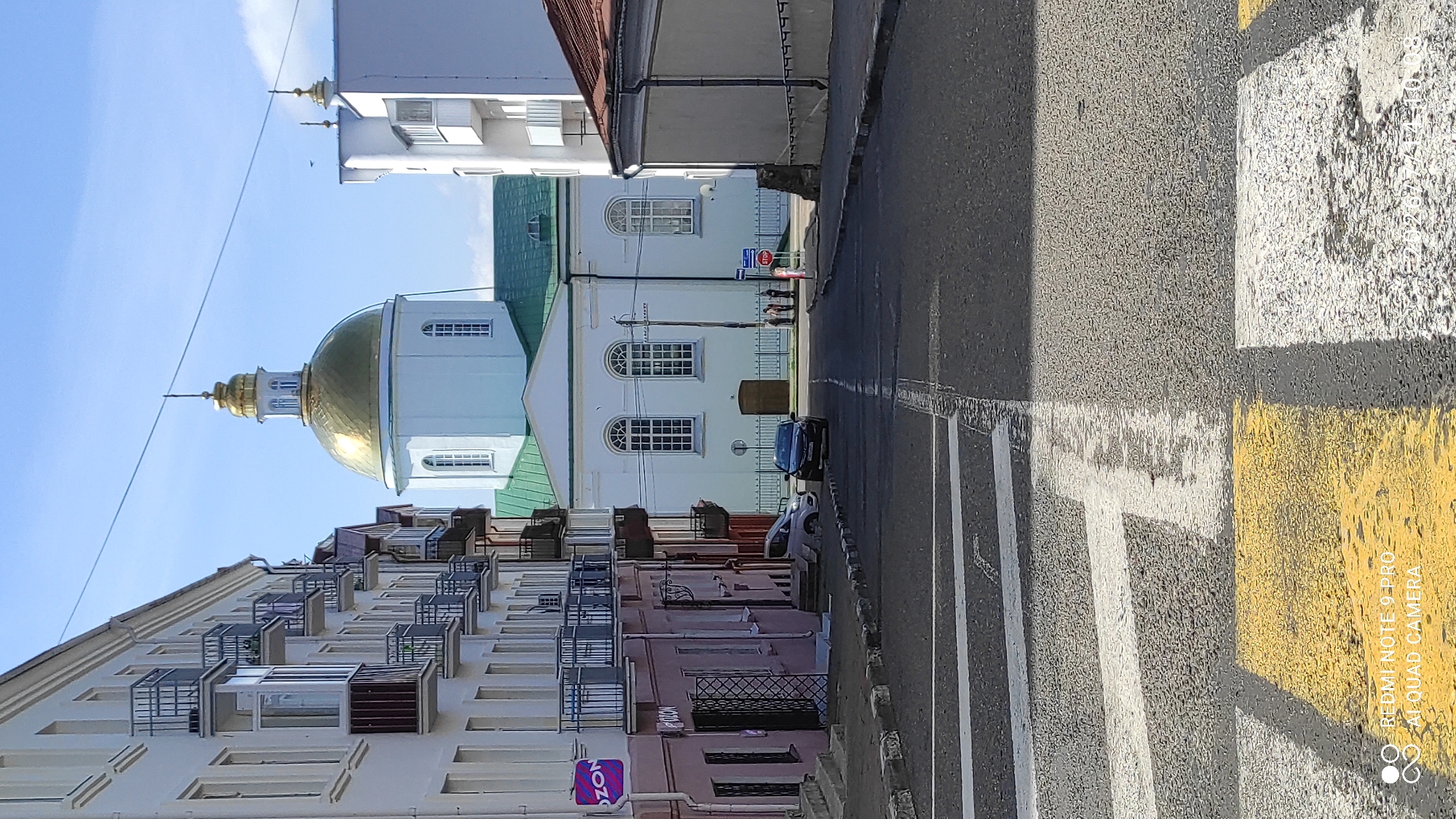 The image depicts a street scene in Polatsk, Belarus, with a prominent white building featuring a golden dome in the background, likely a church or religious institution. The street in the foreground has a pedestrian crossing with yellow and white markings. On the left side of the street, there are multi-story buildings with balconies and shops at ground level, including one with an "OZON" logo. On the right side, there is a lower building with a brown roof. Several cars are parked or driving on the street, and a few people are visible near the building with the golden dome. The sky is clear and blue.