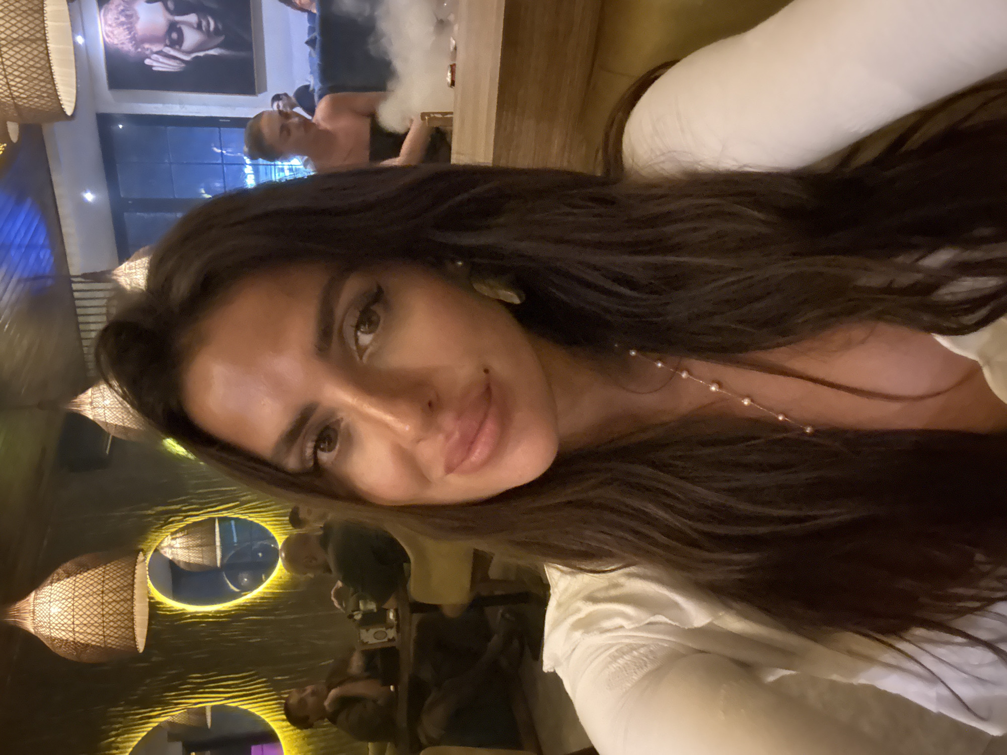The image is a selfie taken by a woman with long, dark hair, wearing a white top and a pearl necklace. She is in a dimly lit indoor setting, possibly a restaurant or lounge. In the background, there are other patrons, tables, chairs, and decorative elements like woven lampshades and mirrors. The overall ambiance appears to be social and relaxed. There is no indication of the city of Kuta Selatan, Indonesia, in the image itself.