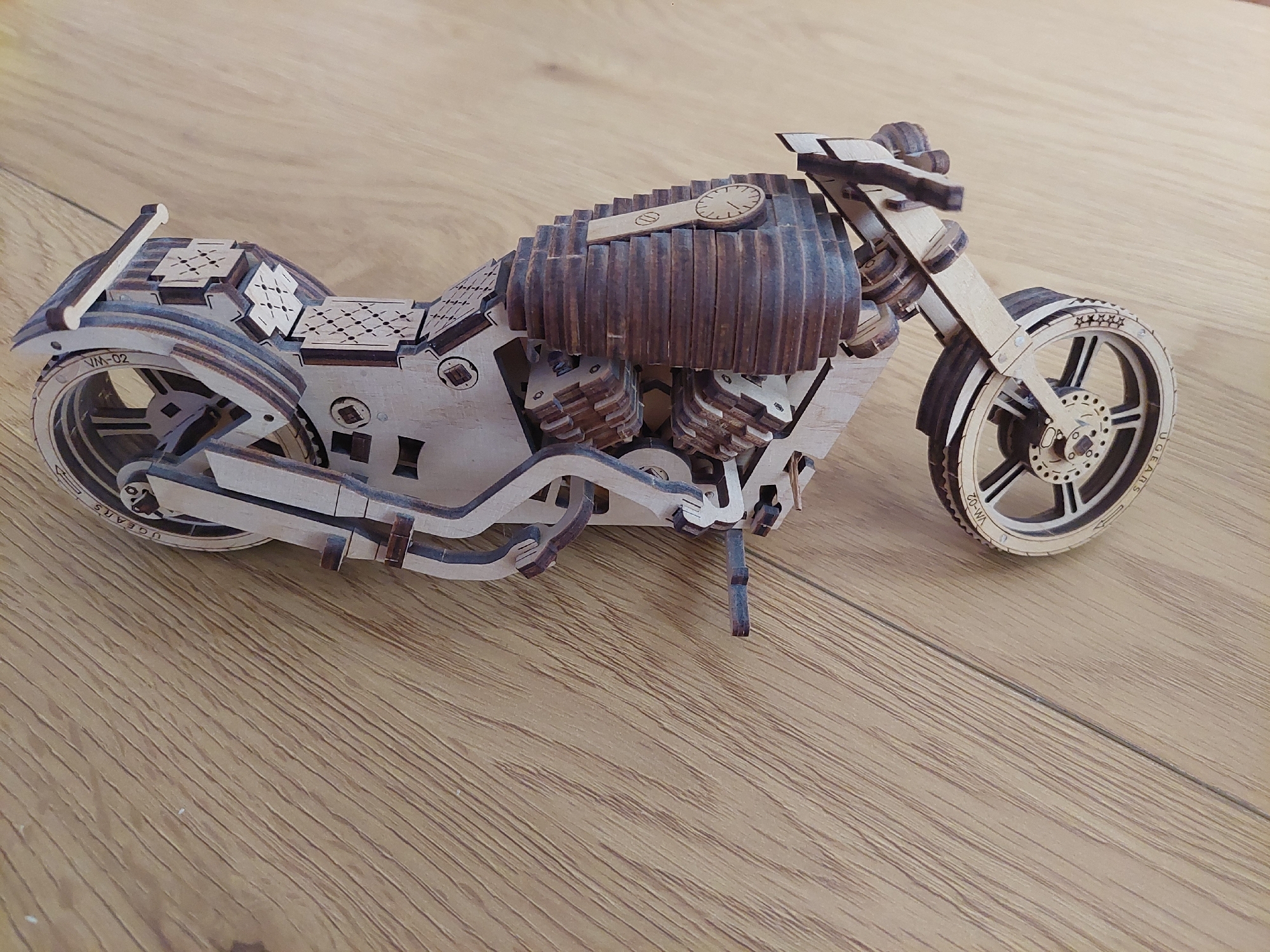 A close-up shot displays a wooden model motorcycle resting on a wooden surface, possibly a table or floor. The motorcycle is intricately assembled from laser-cut wooden pieces. The fuel tank is a dark brown, ribbed construction with a clock face. The front wheel has a five-star detail on the wheel. The tires are marked with the "UGEARS" brand and "VM-02". A kickstand is extended to support the model. The surface beneath has a wood grain pattern. The location in the city of Plovdiv, Bulgaria is not shown in the image.