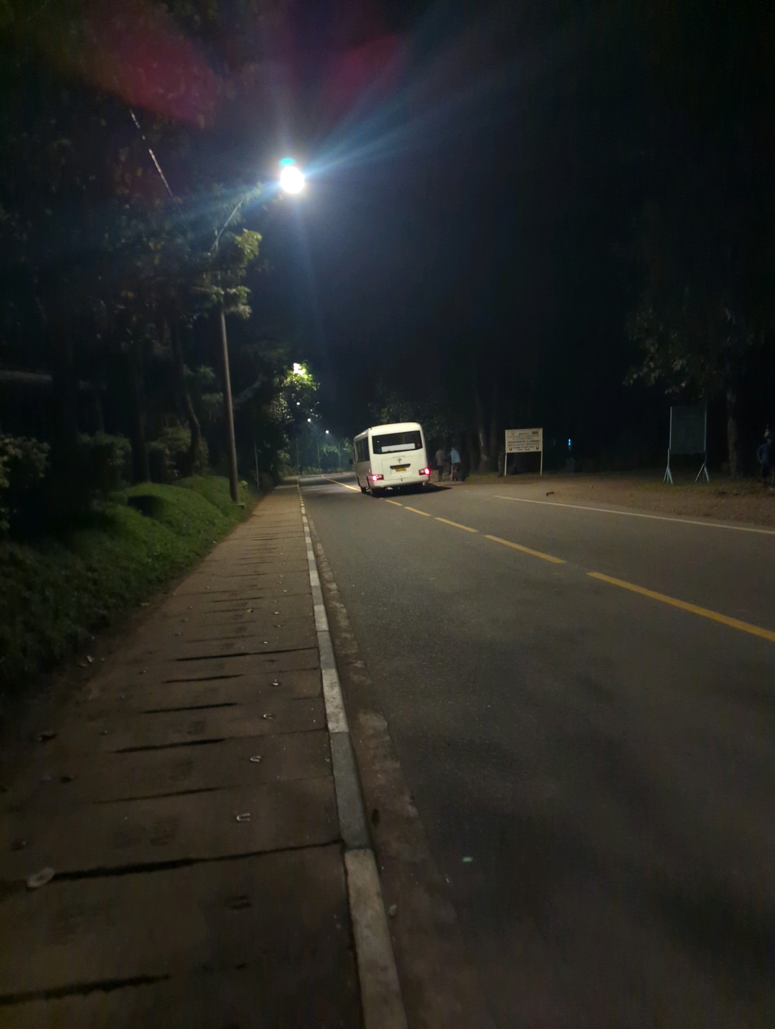 The image depicts a nighttime scene on a paved road in Musanze District, Rwanda. A white bus is parked on the right side of the road, with its brake lights illuminated. Several individuals, appearing to be passengers, are standing near the bus. A sidewalk runs along the left side of the road, bordered by grass and trees. A street light illuminates the scene, casting shadows on the sidewalk. The overall atmosphere is calm and quiet.