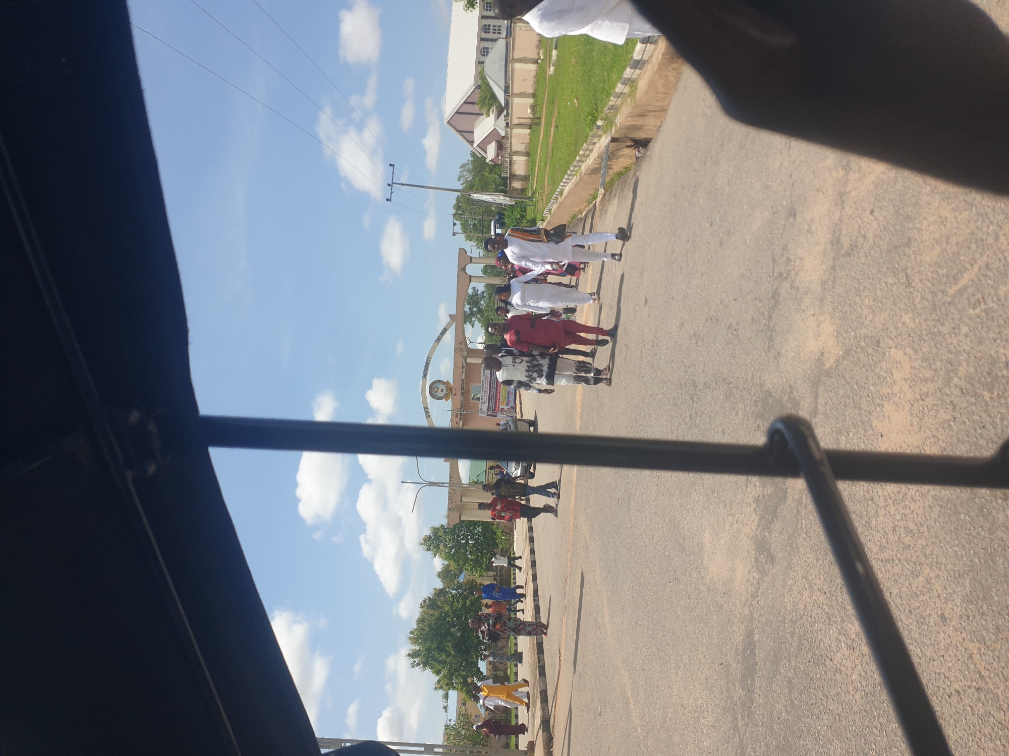 The image, captured from within a vehicle looking forward and slightly right, depicts a sunny daytime scene on an asphalt road in Jalingo, Nigeria, specifically at the entrance to Taraba State University.
In the background, a large archway structure, identifiable as a university gate, displays the text "STATE UNIVERSITY, JALINGO" and partially visible "TARABA STATE UNIVERSITY" on its facade. A circular emblem or clock is centered above the archway. To the right of the gate, a prominent building with a reddish-brown gabled roof and white walls stands amidst green grass and utility poles with overhead wires. Lush green trees are visible behind the archway and to the left. The sky is clear blue with scattered white cumulus clouds.
Numerous individuals are present on and alongside the road. On the left side, several people, some in yellow/orange garments, walk away from the viewer. In the mid-ground, a diverse group of people walks in various directions. A woman in a patterned long dress stands prominently in the center-left. Further right, a group of individuals, many dressed in traditional Nigerian attire including white and patterned fabrics, walk towards the camera. Two men, one in a red shirt and the other in a darker top, walk near the right edge of the road, also approaching. A person in white clothing is partially visible in the far right foreground. The road itself is paved, with some light-colored patches suggesting dust or dirt, and a low concrete curb borders a grassy verge on the right.