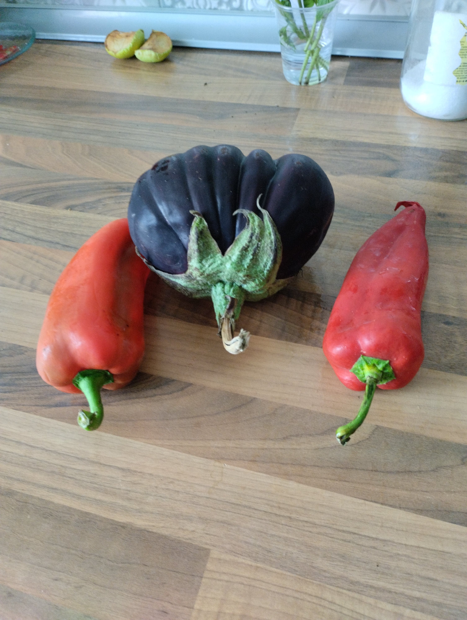 In the city of Mezitli, Turkey, on a wooden kitchen counter, there are three vegetables: a large, dark purple eggplant in the center, and two bright red bell peppers, one to the left and one to the right of the eggplant. The eggplant has a prominent green calyx. The bell pepper on the left is positioned with its stem facing down and to the left, while the bell pepper on the right has its stem pointing upwards and to the right. In the blurred background, to the left, there are two pieces of sliced green apple on the counter. To the right of the apples, there is a clear glass of water with green stems of what appears to be mint or basil. Further to the right in the background is a white plastic container with a label.