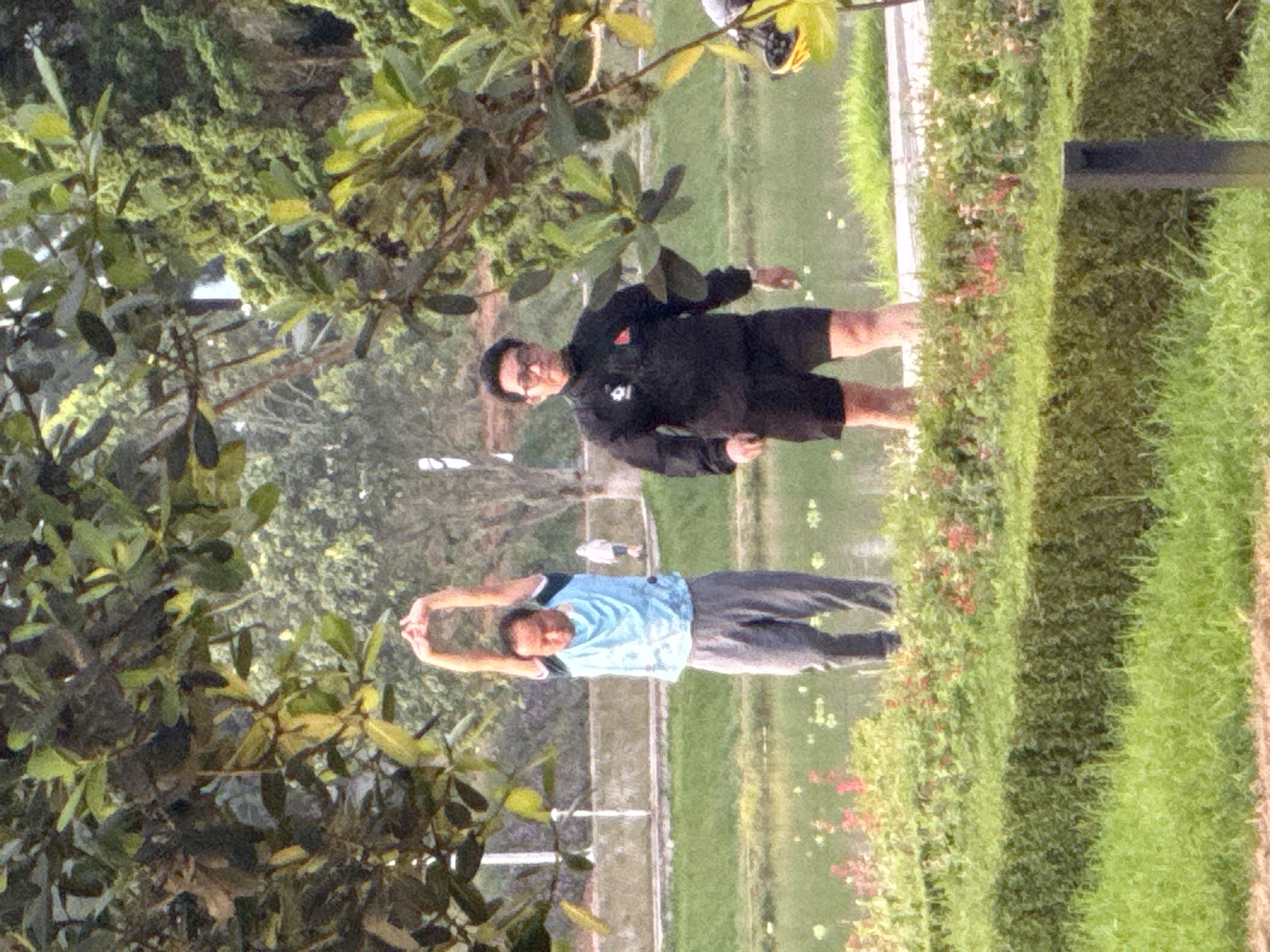 Two men are in a park with a body of water and lush greenery. The man on the left is wearing a blue t-shirt and grey sweatpants, with his arms raised above his head. The man on the right is wearing a black jacket and black shorts, with glasses. He appears to be walking. In the background, there are trees, a walking path, and another person in the distance. The scene suggests a recreational setting in a city. The location is identified as Phường Xuân Hương - Đà Lạt, Vietnam.