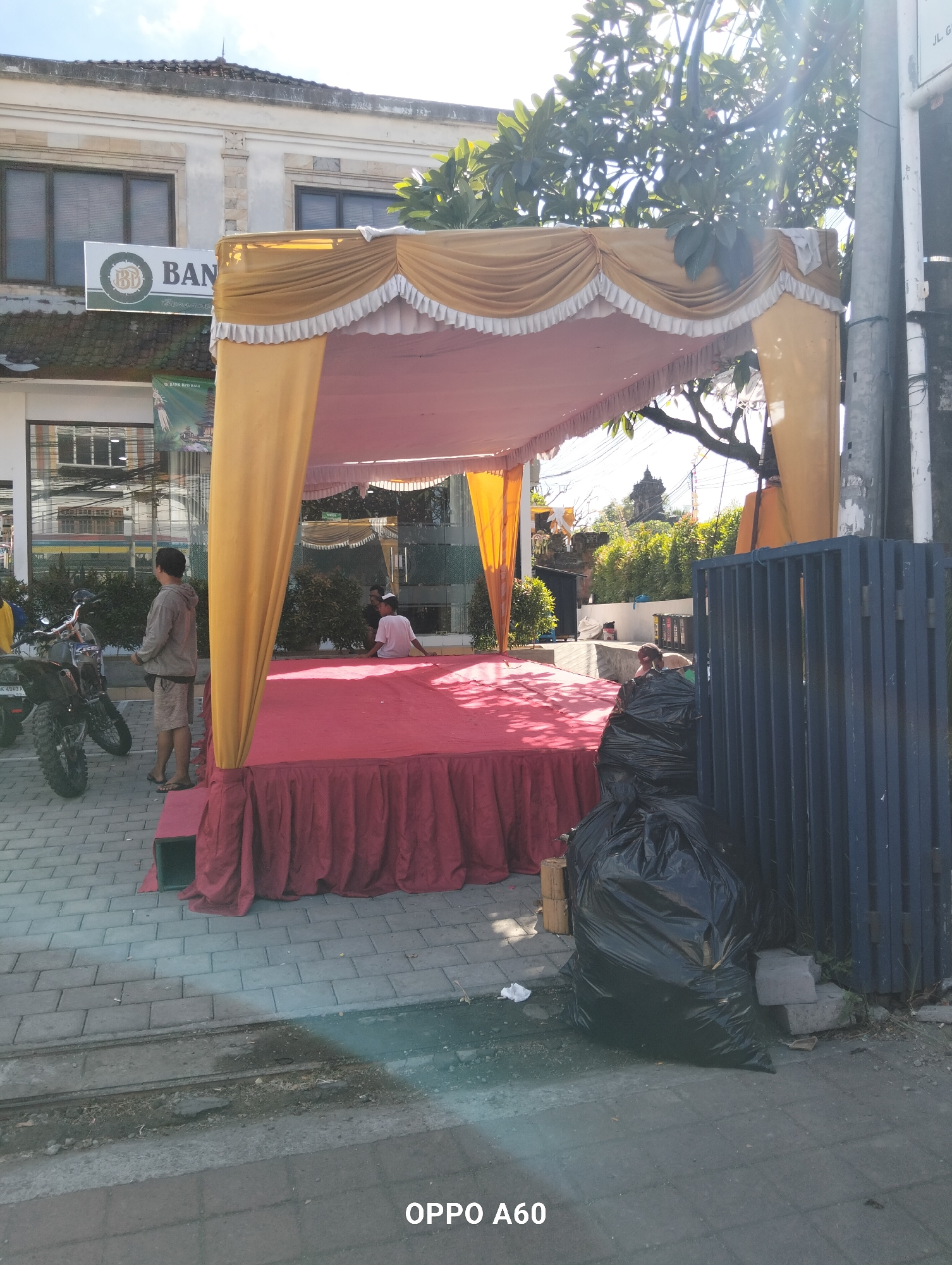 An outdoor scene in North Kuta, Indonesia features a red-skirted stage under a yellow canopy. Two men are present: one stands to the left of the stage, facing away from the camera, wearing a grey hoodie and shorts. Another man sits on the stage, facing left and away from the camera, wearing a white t-shirt. A black garbage bag is placed to the right of the stage. In the background, a building with a "BANK" sign is visible, along with trees and a blue metal fence. Motorcycles are parked to the left of the stage. The ground is paved with interlocking bricks.