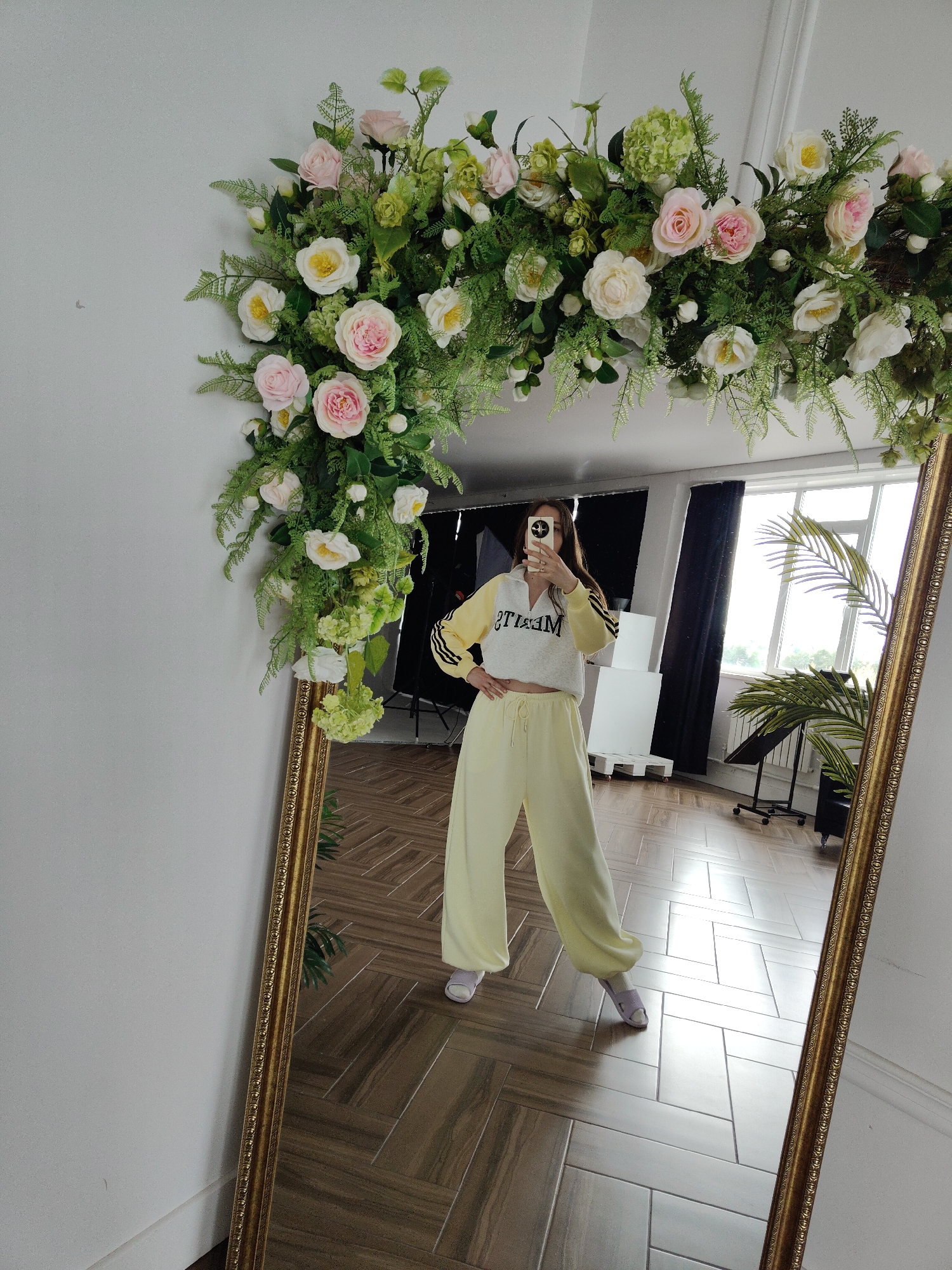 A full shot captures a person standing in front of a mirror with a floral garland attached to the top. The person is reflected in the mirror, holding a smartphone up to take the picture. The individual is wearing a yellow and white tracksuit, with the sweatshirt cropped to expose the midriff, and yellow pants cinched at the waist with a drawstring. The person is also wearing lavender sandals. The floral garland consists of pink and white roses, interspersed with green foliage.
The background shows a room with a tiled floor and a large window letting in natural light. A green plant can be seen near the window, along with some white and black studio equipment. The location appears to be a studio setting.