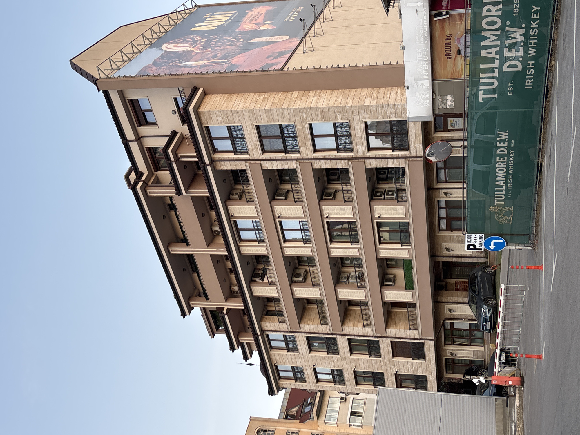 A six-story modern building with a tiered facade is shown on a bright, clear day in Sofia, Bulgaria. The building is primarily beige with brown accents and features multiple balconies with black metal railings. Many windows with dark frames are visible. A large billboard advertising "Nuii" ice cream is attached to the right side of the building.
In front of the building, a green mesh fence with large "Tullamore D.E.W." branding is erected. A street with two parked cars, a gray SUV and a black car, is visible in the foreground. A red and white retractable barrier is positioned across the street, suggesting an entrance or exit to a parking area. A blue sign with an arrow pointing left and the word "PARKING" in Cyrillic script is mounted on a pole to the right of the barrier. The sky is a clear, light blue.