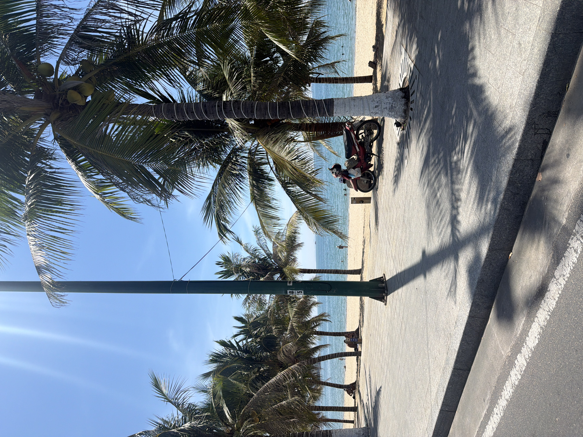 The image depicts a sunny day at a beach in Nha Trang Ward, Vietnam. In the foreground, a paved walkway with a curb and a street with a white line are visible. Palm trees with coconuts cast shadows on the walkway. A green lamppost stands prominently on the walkway, with wires extending from it. To the right, a red motorcycle is parked on the walkway, with a white bag hanging from it. The background features a sandy beach with several people relaxing, and the ocean with calm water. The sky is clear and blue with visible sun rays. The scene suggests a typical beach day with leisure activities.