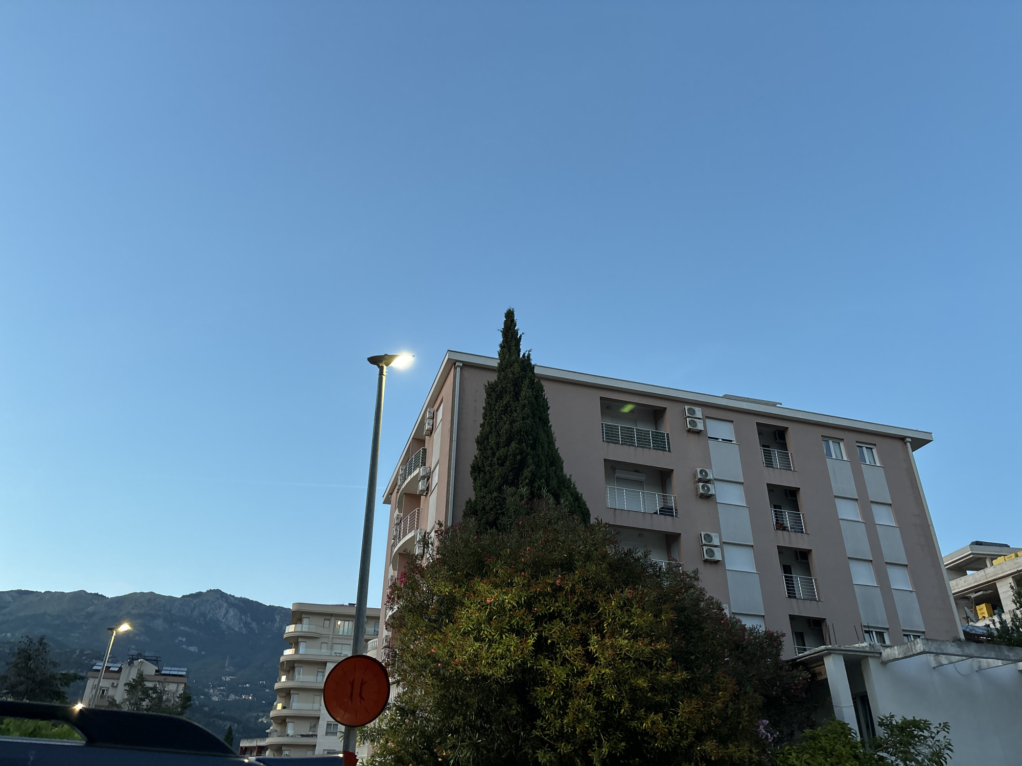 The image depicts an exterior scene in Budva, Montenegro, under a clear blue sky. In the foreground, a dark-colored vehicle is partially visible. A street lamp with its light on stands to the left of a tall, slender cypress tree. To the right of the cypress tree is a multi-story building with a light-colored facade, featuring balconies with railings and several air conditioning units mounted on the exterior walls. A circular orange traffic sign with a red border and white arrows pointing in opposite directions is positioned in front of the cypress tree and the building. In the middle ground, another building with a similar architectural style is visible, along with lush green foliage. Further in the background, a range of mountains with a few scattered buildings on their slopes is prominent. The overall scene suggests an urban environment with natural elements.