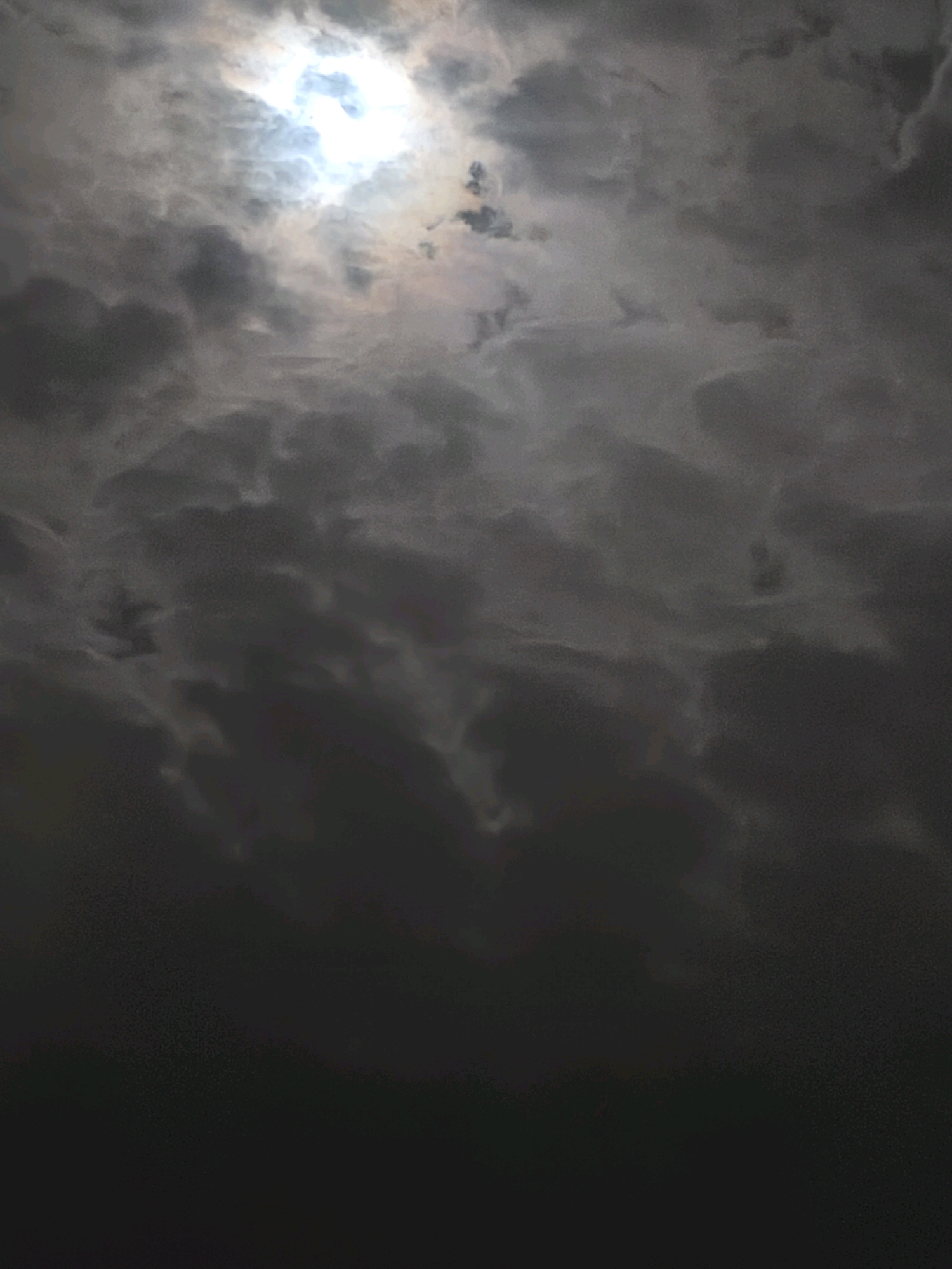 The image depicts a dramatic sky over Passignano sul Trasimeno, Italy. A bright, diffused light source, likely the sun obscured by clouds, is visible in the upper left quadrant. The sky is filled with dense, dark, and turbulent clouds that create a somber and moody atmosphere. The lower portion of the image fades into complete darkness, suggesting a landscape or foreground that is not illuminated. No people, objects, or specific landmarks are discernible in the image, focusing solely on the atmospheric conditions of the sky.