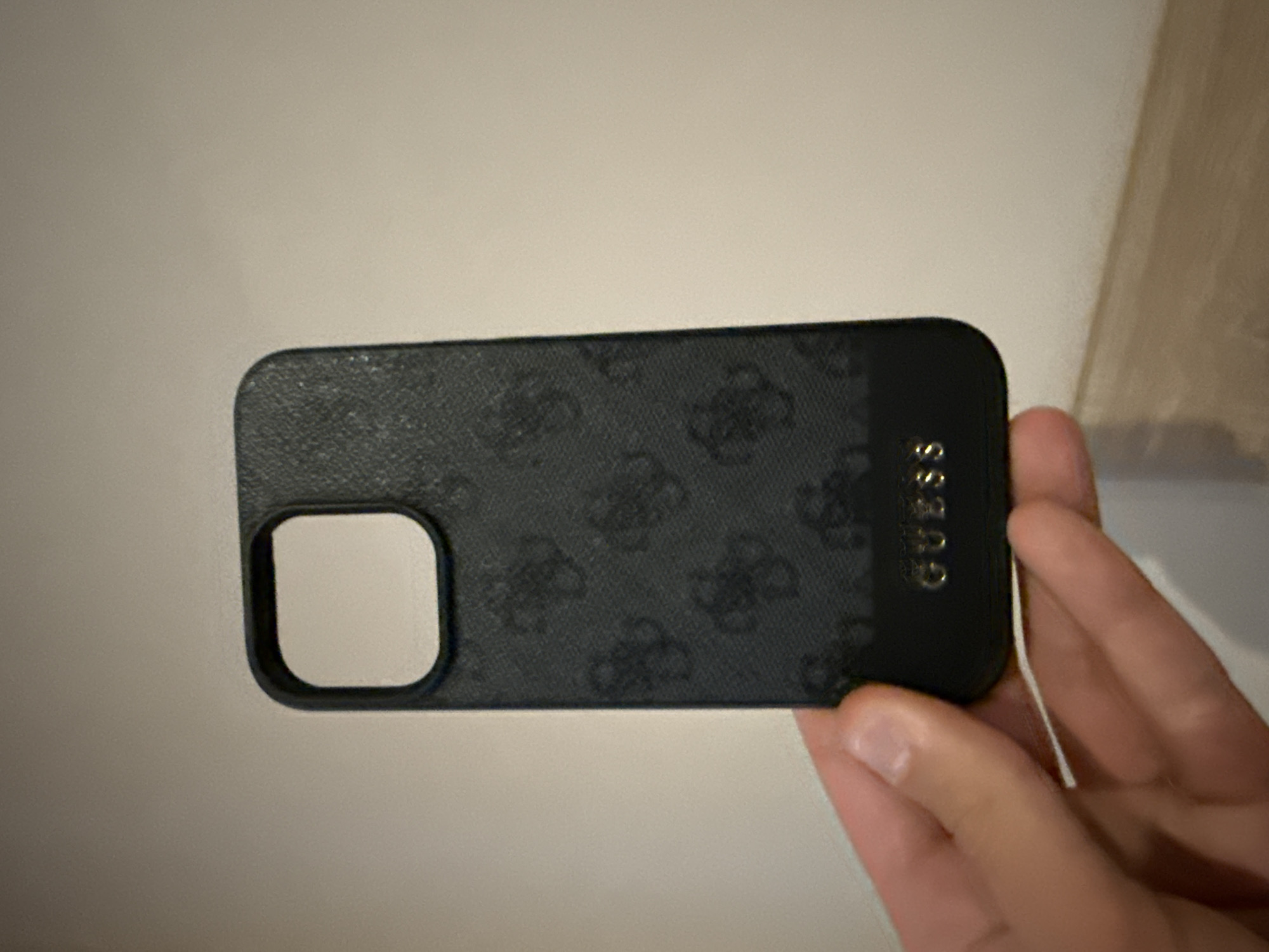 A close-up shot shows a black Guess-branded phone case held in a person's hand. The case features a repeated Guess logo pattern. The background is a plain white surface, potentially a wall or table. The photo does not contain information that can be used to infer any location, including Lovech, Bulgaria.
