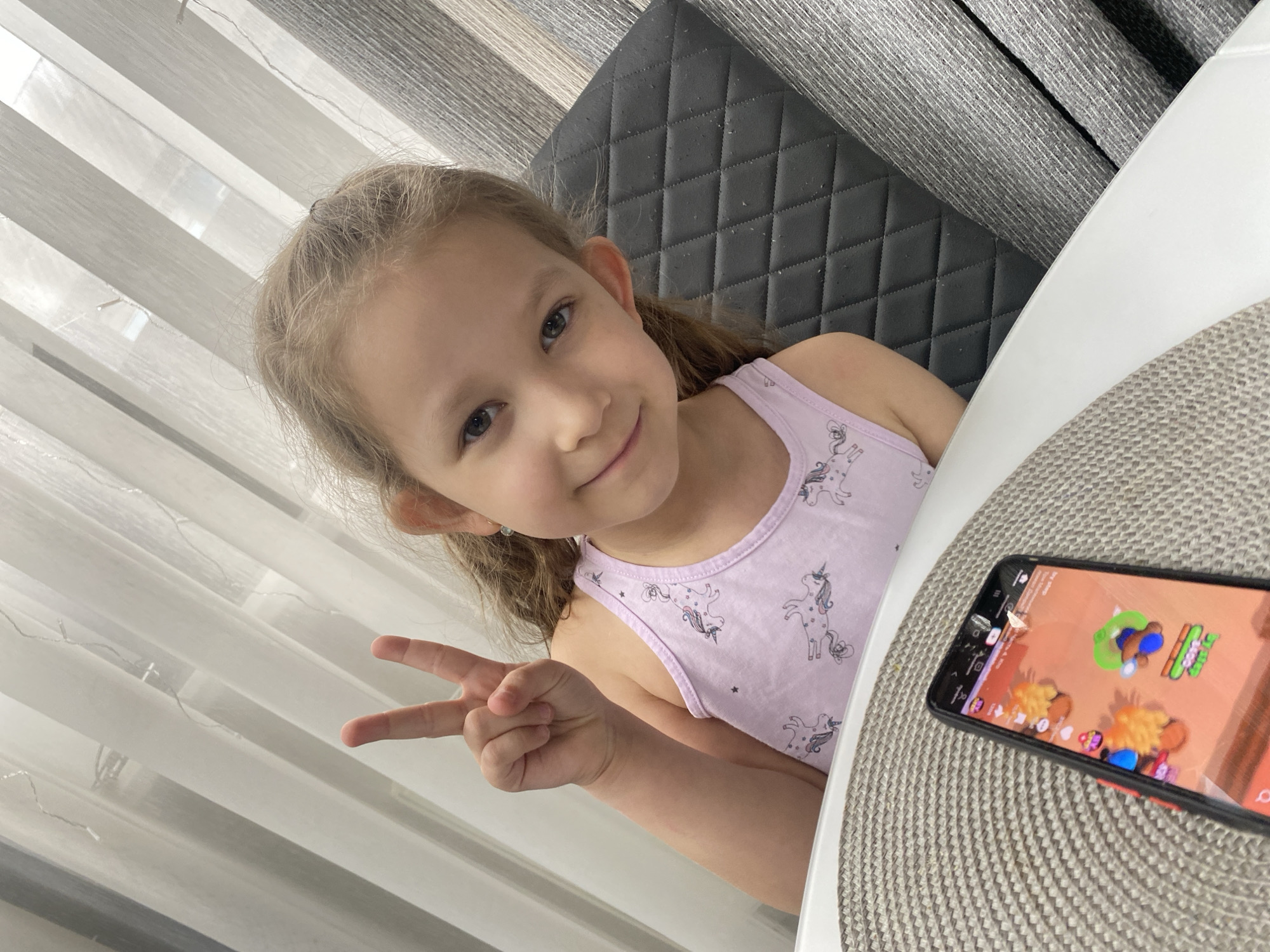 A young girl with light brown hair and a happy expression is seated indoors, likely at a table, in Izhevsk, Russia. She is looking directly at the camera and making a peace sign with her left hand. She wears a pink tank top adorned with unicorn patterns. Behind her, a dark, quilted chair back is visible, and further back, horizontal gray and white striped window blinds suggest a daytime setting with natural light. On the light-colored table to her right, a smartphone rests on a round, textured placemat. The phone screen displays a colorful game with various animated characters and elements, though specific text on the screen is illegible due to pixelation and distance. The overall mood is cheerful and relaxed.
