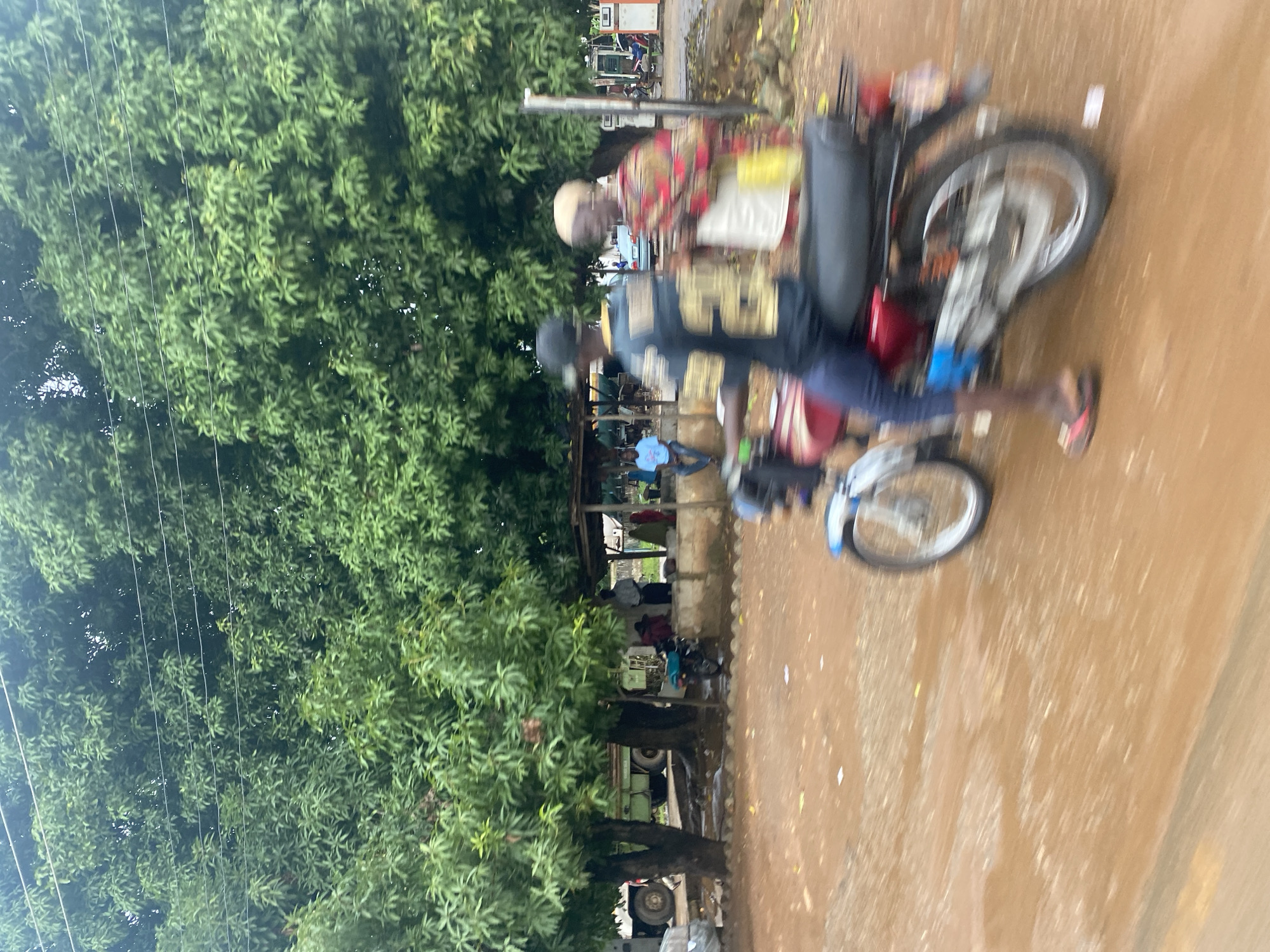 A motion-blurred, eye-level, outdoor shot captures a street scene in Keffi, Nigeria, with lush green trees dominating the background and framing the central activity. In the foreground, a person on a red motorcycle is moving from left to right. The rider, wearing a dark baseball cap, a dark blue shirt with gold lettering and numbers, and dark pants, is partially obscured. Their legs, one with a red sandal, are visible. To the right of the motorcycle, another person is walking away from the camera, wearing a brightly colored, patterned top and a white and yellow garment.
Behind the motorcycle, a rustic structure with a thatched or corrugated roof provides shade. Beneath it, several individuals are present; some appear seated, while others stand. One person, wearing a blue shirt, is sitting on a low wall. A parked motorcycle is visible under the structure. Further back, a green truck is parked to the left, and in the distance, what appears to be a gas station with pumps and a white structure can be seen. The ground is a reddish-brown, appearing somewhat damp, with scattered white debris. The overall impression is of a bustling, everyday street scene in a tropical or sub-tropical environment.