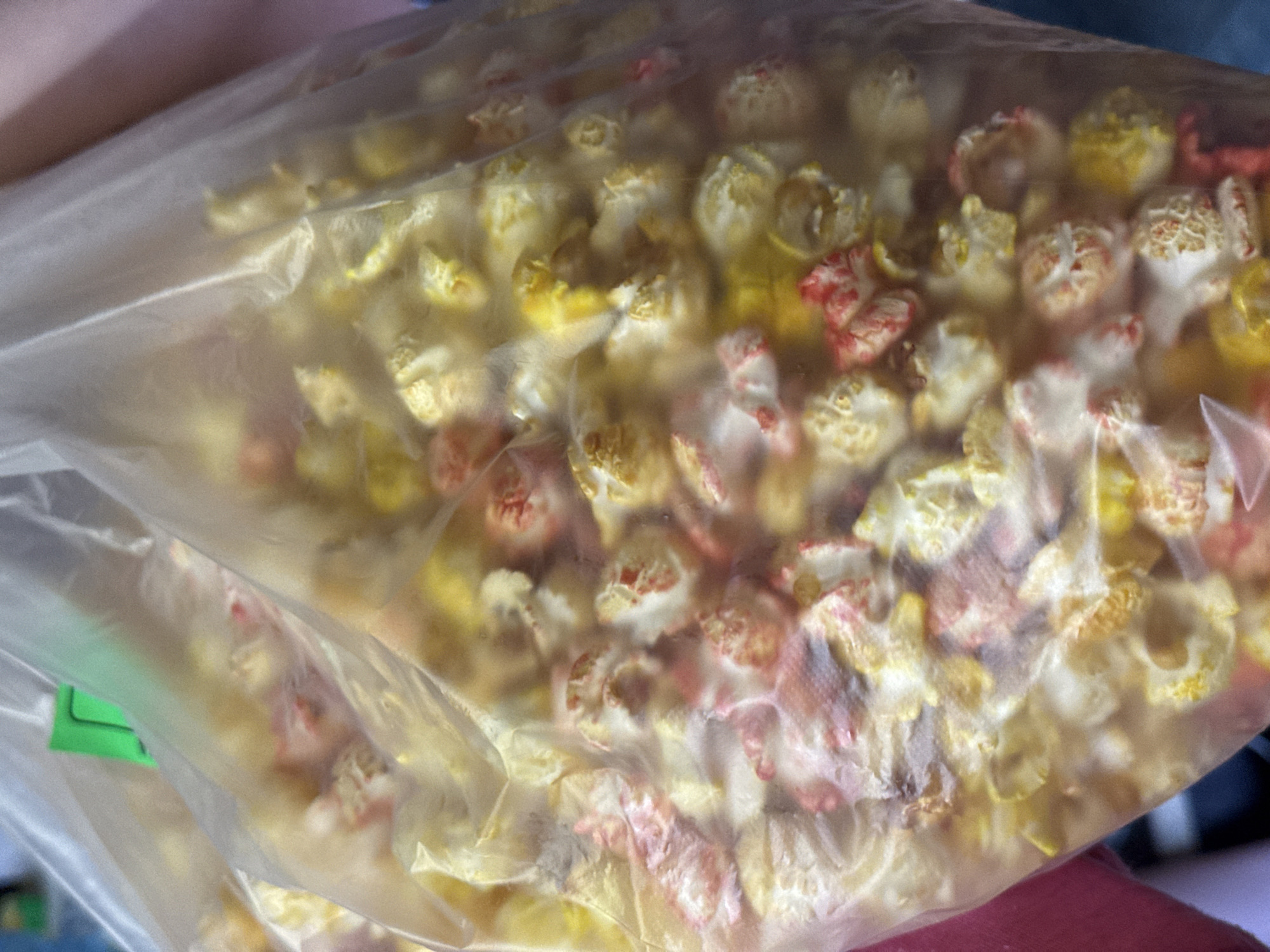 A close-up, eye-level shot captures a large, clear plastic bag filled with multicolored popcorn. The popcorn pieces are predominantly yellow, red, and white, suggesting different flavor coatings. The plastic bag exhibits a crinkled texture and reflections from ambient light. A bright green plastic clip or tag is visible at the top left edge of the bag, securing or identifying it. The bag appears to be held, with a blurred reddish-pink fabric visible on the left side and a darker, indistinct background element on the right. The focus is sharp on the popcorn within the bag, which is densely packed. The scene is located in Unknown, Unknown.
