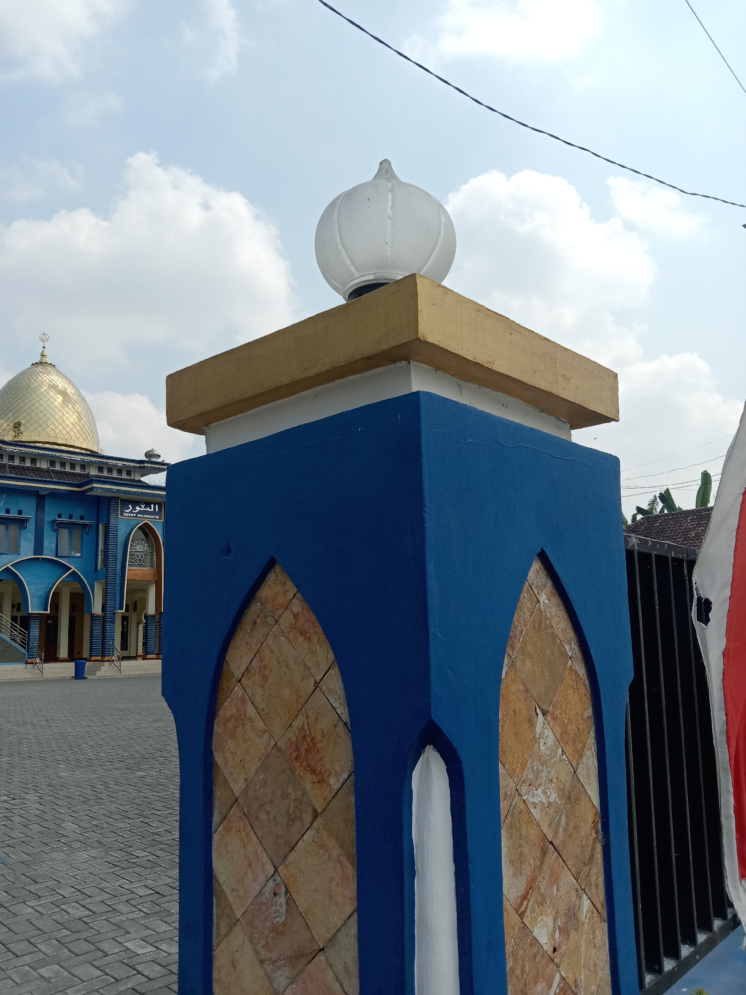 The image shows a detail of a blue pillar in the foreground, topped with a golden rectangular structure and a white, dome-shaped light fixture. The pillar has an arched opening filled with diamond-patterned brown tiles. In the background, a blue mosque with a golden dome is visible. The mosque features Arabic script above an arched entrance. The ground in front of the mosque is paved with gray interlocking stones. The scene is set under a blue sky with scattered white clouds. The location is the Al-Munawwar Sepat Mojoroto Mosque in Sidobandung, Indonesia.