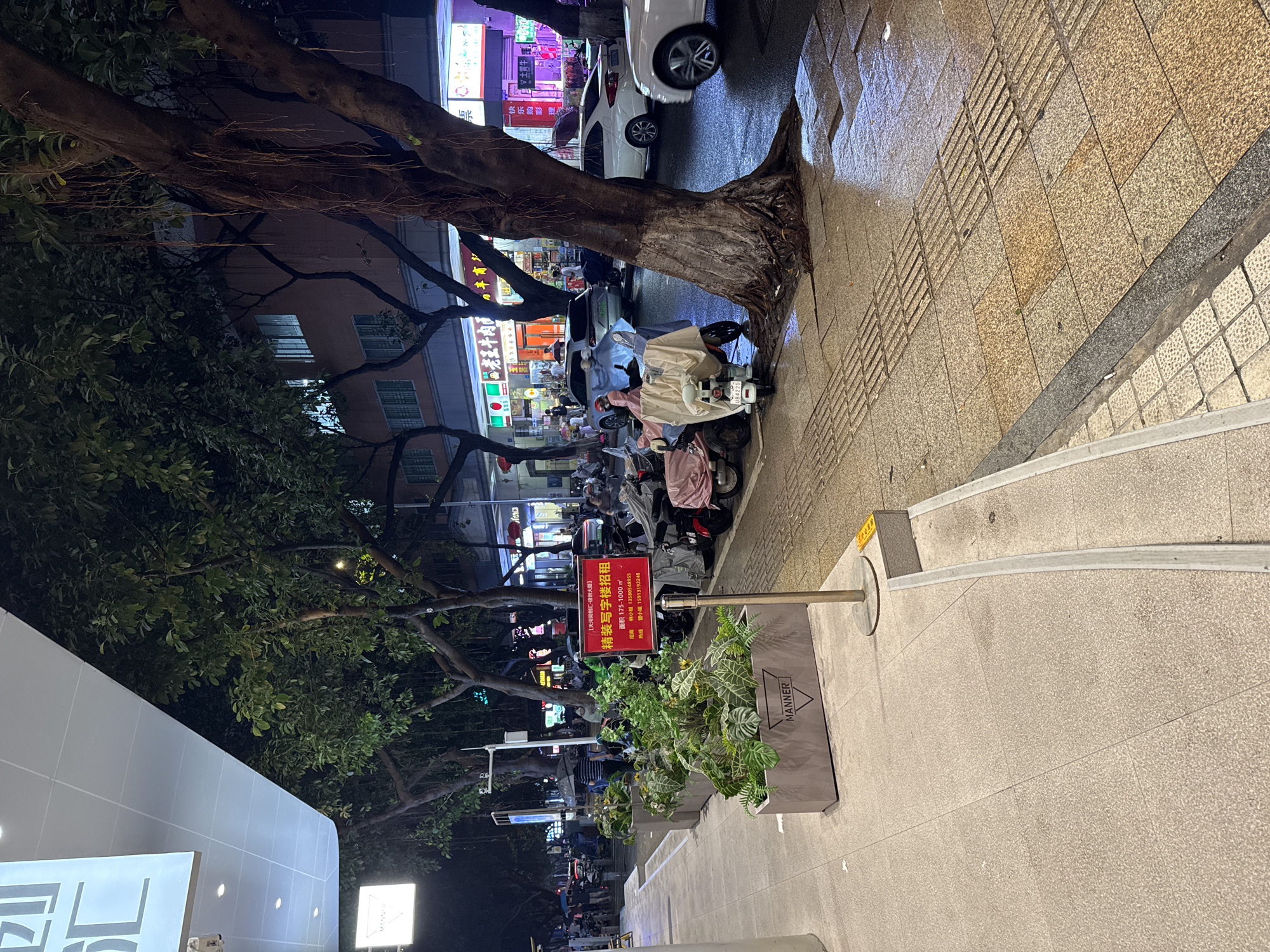 Here is a description of the image:
The image depicts a street scene at night in Tianhe District, China, specifically near the "Tianhe Tongchuanghui - Xin Chuang Building" area. The street is lined with trees, and the sidewalks are paved with textured tiles. A large tree with exposed roots is visible on the right side of the image, growing close to the edge of the sidewalk. Cars and scooters are parked along the street.
A sign written in Chinese advertises fully furnished office spaces for rent. A rectangular planter with the word "MANNER" on it is also visible next to the rental sign. Further down the street, the storefronts display signage in Chinese. Overall, the scene captures a typical urban environment at night, with a mix of commercial activity and parked vehicles.