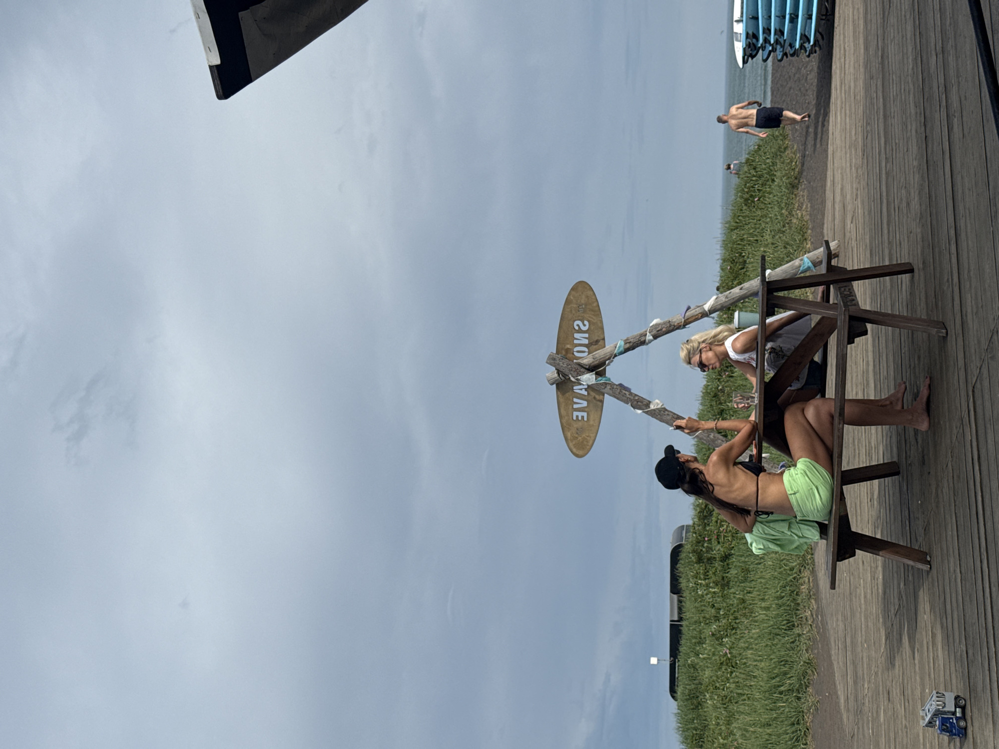 Two women are seated at a picnic table under a triangular wooden structure with a surfboard-shaped sign that reads "SNOWAVE" in reverse. The woman on the left wears a black bikini top, neon green shorts, and a black baseball cap. She is holding a cigarette and appears to be talking to the woman on the right. The woman on the right wears a white tank top and sunglasses and is looking to her left. A green towel is draped over the back of the bench on the left. On the wooden boardwalk in front of the table, a small blue toy truck is visible. In the background, there is a grassy hill, a beach, and the ocean. A man in dark shorts is walking away from the camera on the beach. Stacks of blue surfboards are leaning against a black structure on the right side of the image. The sky is overcast and gray. The scene is located in Petropavlovsk-Kamchatsky, Russia.