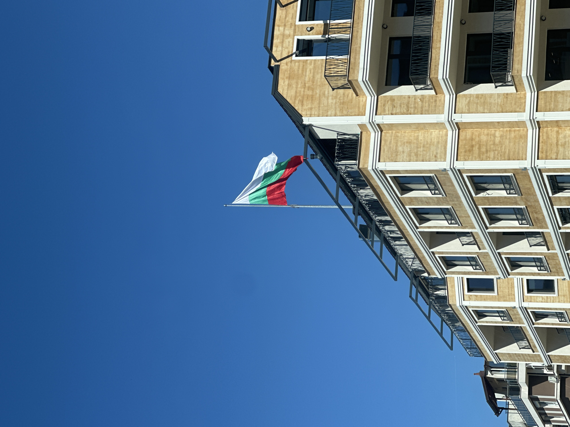 The image shows a building with a Bulgarian flag flying from it, set against a clear blue sky. The building appears to be a multi-story apartment or commercial building with balconies and a facade that has a pattern of recessed rectangular panels, framed by white trim. The flag, displaying the characteristic white, green, and red horizontal stripes, is attached to a bracket extending from the building. The image is rotated. The location is likely in Sofia, Bulgaria, due to the presence of the Bulgarian flag.