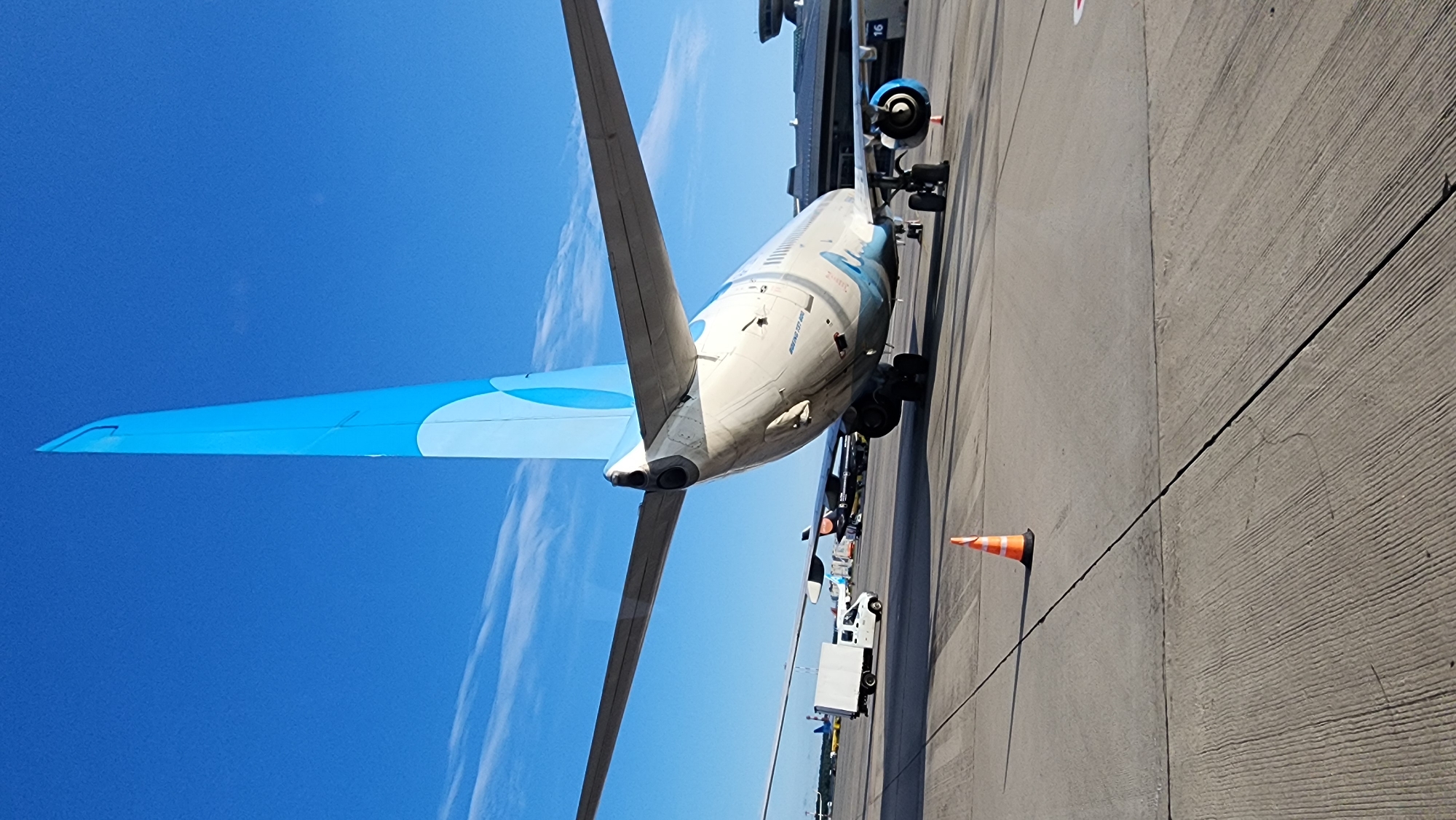 The image shows the tail section of a Boeing 737-800 aircraft with the airline livery of "Azur Air" which is a Russian charter airline. The aircraft is parked on the tarmac at Vnukovo International Airport in Moscow, Russia. The tail is painted white with a light blue curved design. The aircraft is positioned facing away from the viewer, with its wings extending to the sides. In the background, other aircraft and airport service vehicles are visible, along with a terminal building. The sky is clear blue with some scattered white clouds. The tarmac is paved with concrete and shows tire marks and cracks. An orange traffic cone is placed on the tarmac in the foreground. The lighting suggests it is daytime, likely late afternoon or early morning, given the long shadow cast by the cone. There are no people visible in the image.