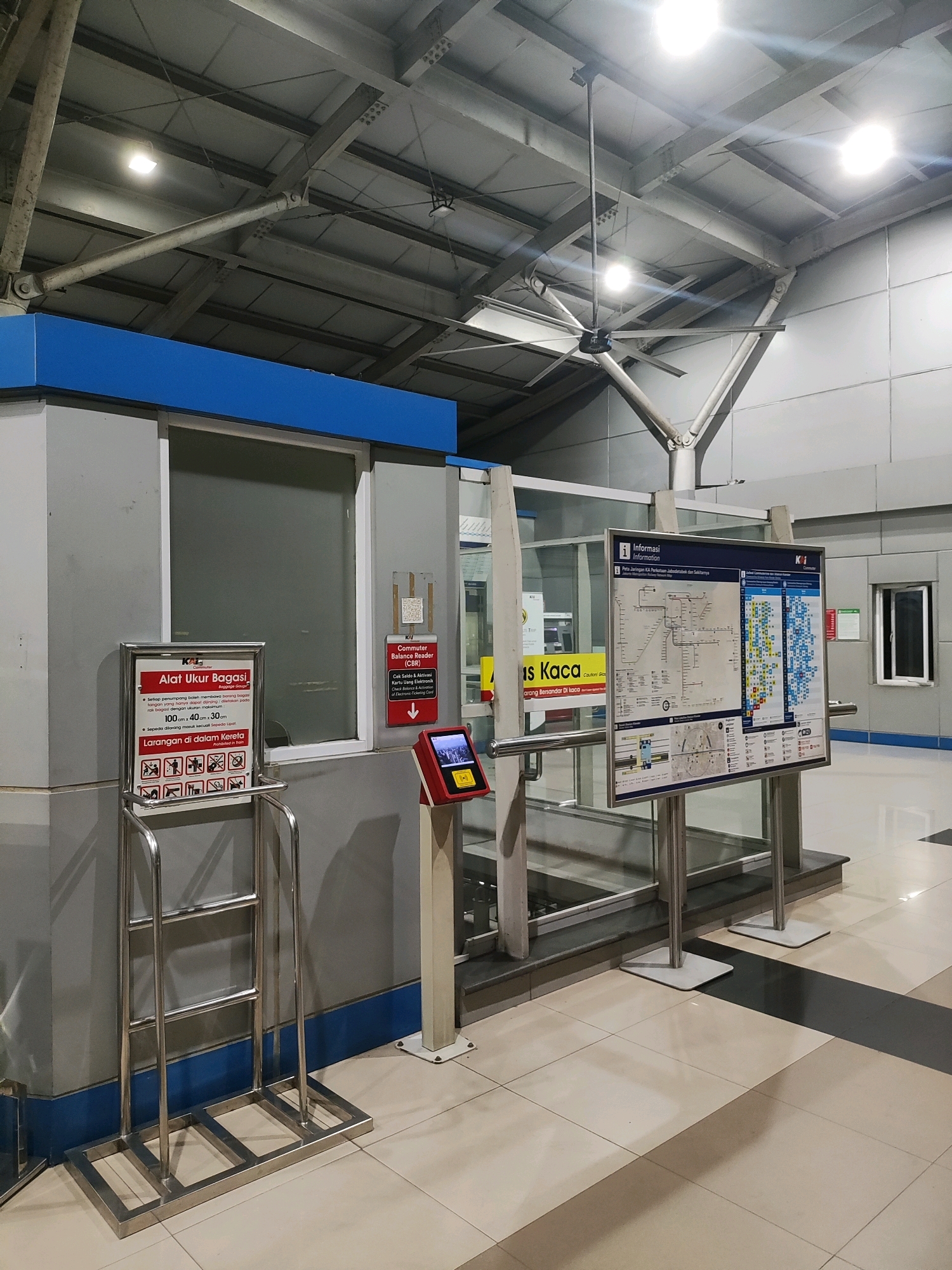 The image depicts the interior of a train station concourse in East Jakarta, Indonesia. The scene is well-lit by artificial lights and features a large industrial-style ceiling fan. In the foreground, a metallic rack designed to hold luggage or bicycles stands on a tiled floor. To the left, a ticket or information booth is visible, with a sign detailing luggage size restrictions and prohibited items, along with a smaller sign about bicycle transport. A red and silver machine with a touchscreen display is mounted on a stand next to the booth. To the right, a large information board displays a map of the train network and other relevant information. The overall environment suggests a functional and modern public transportation hub. There are no people visible in the image.