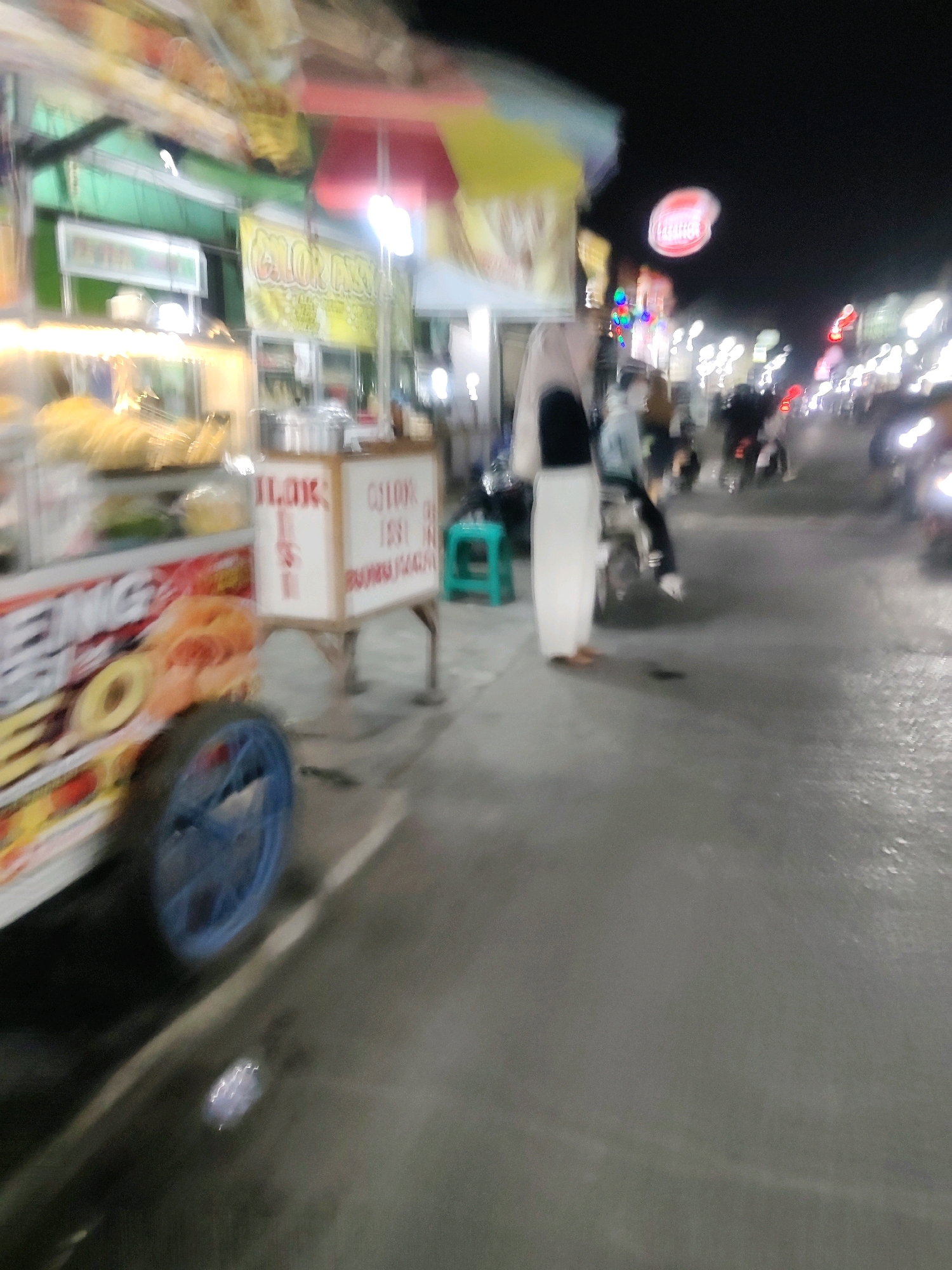 The image depicts a night scene on a street in Mekarmukti, Indonesia, likely a commercial area with food vendors and passing traffic. In the foreground, a blurred food cart is visible, displaying various items and signage, with a blue wheel prominent on the left. To the right of the cart, a white box with red lettering stands on legs. A person wearing a white garment and a dark top is standing near the cart, facing away from the camera. Behind them, several motorcycles are visible, some with riders, moving along the wet asphalt street. The background is filled with the glow of streetlights, vendor lights, and vehicle headlights, creating a bustling and somewhat chaotic atmosphere. The overall impression is one of a lively street food scene at night.